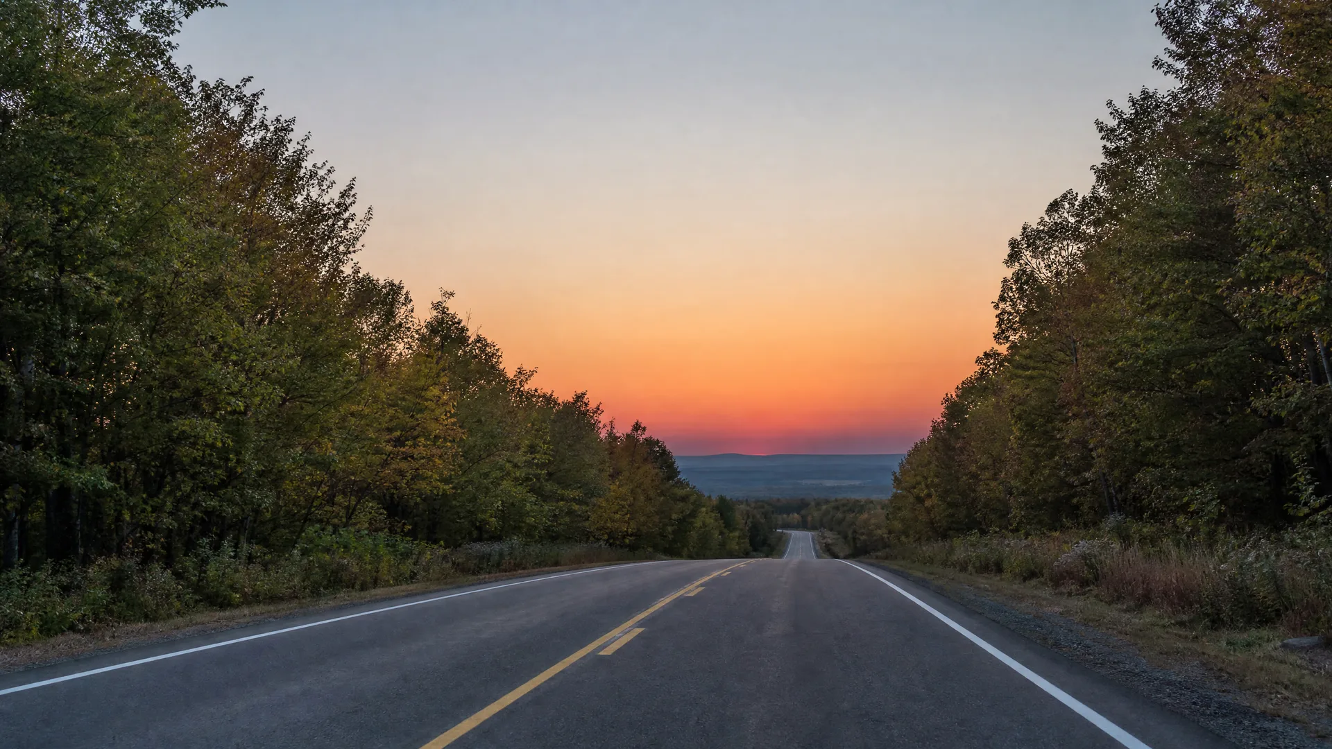 A two-lane paved road with a double yellow center line and white edge lines runs toward a distant tree-lined horizon under an orange and blue sunset sky, flanked by dense deciduous trees.