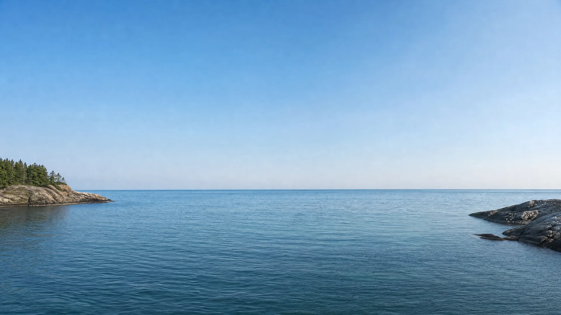 Calm blue water under a clear sky, with a conifer-topped rocky point on the left and bare rock outcrops on the right.