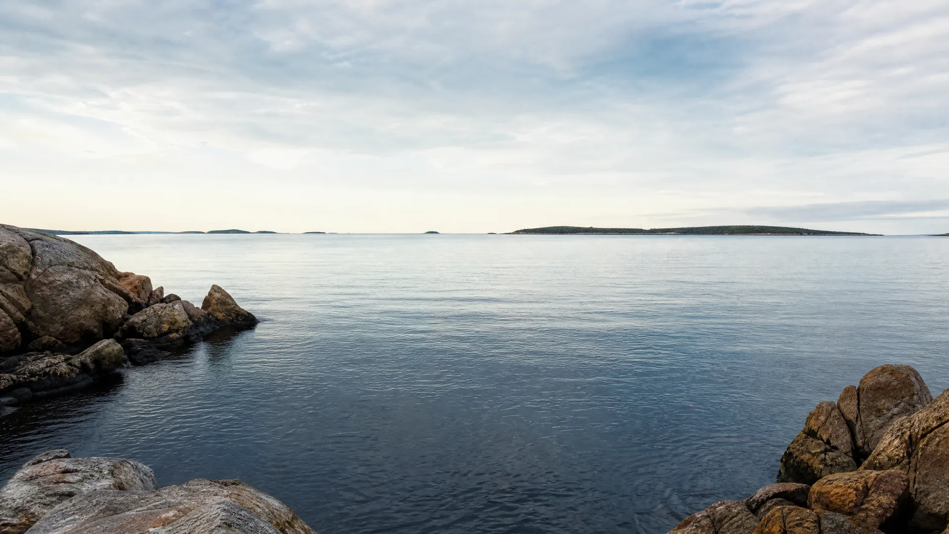 Calm sea seen from a rocky shoreline, with low tree-covered islands on the horizon under a pale, lightly clouded sky.