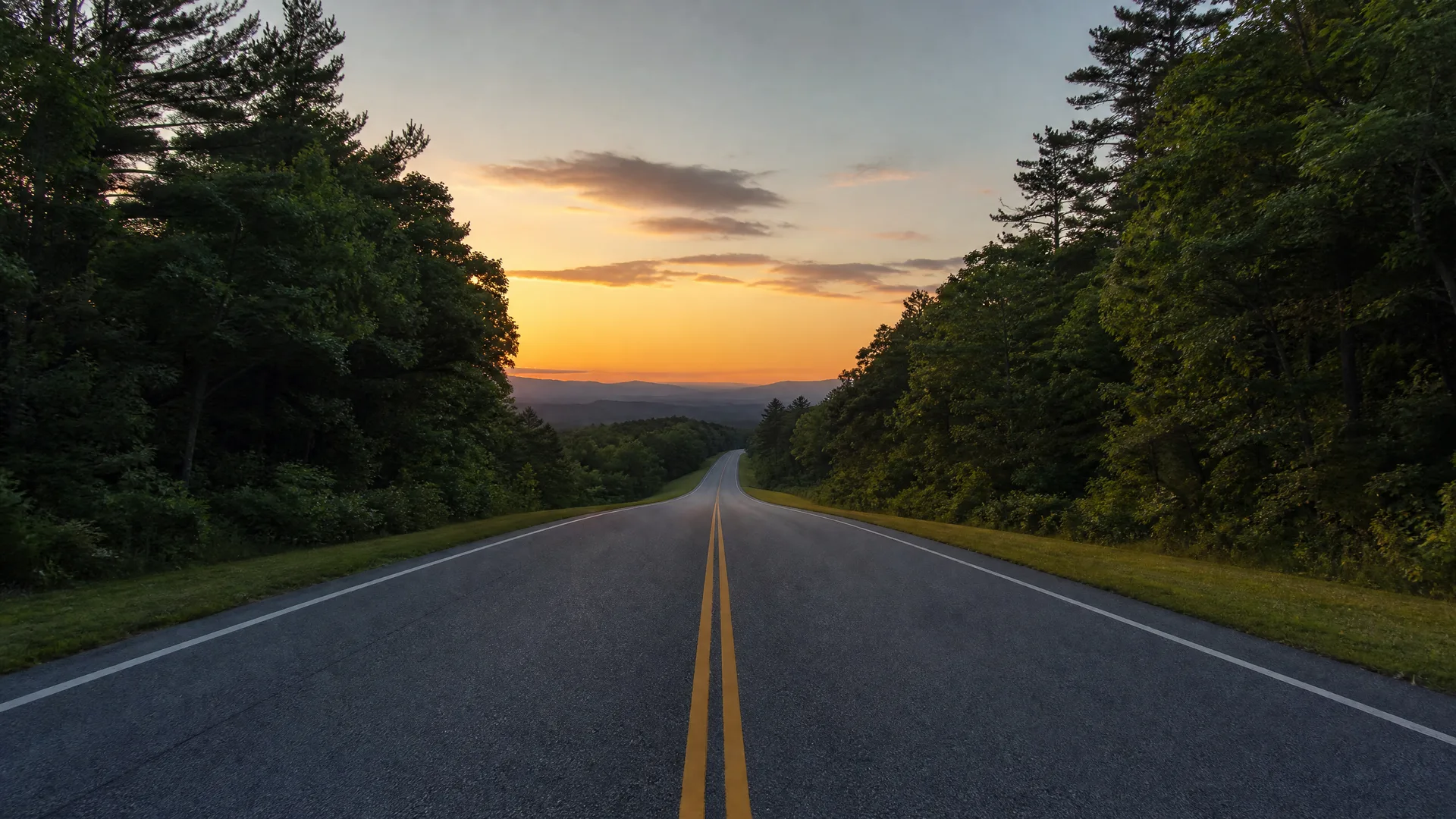 A two-lane asphalt road with double yellow center lines runs toward distant hills, flanked by grassy shoulders and dense trees under an orange sunset sky.