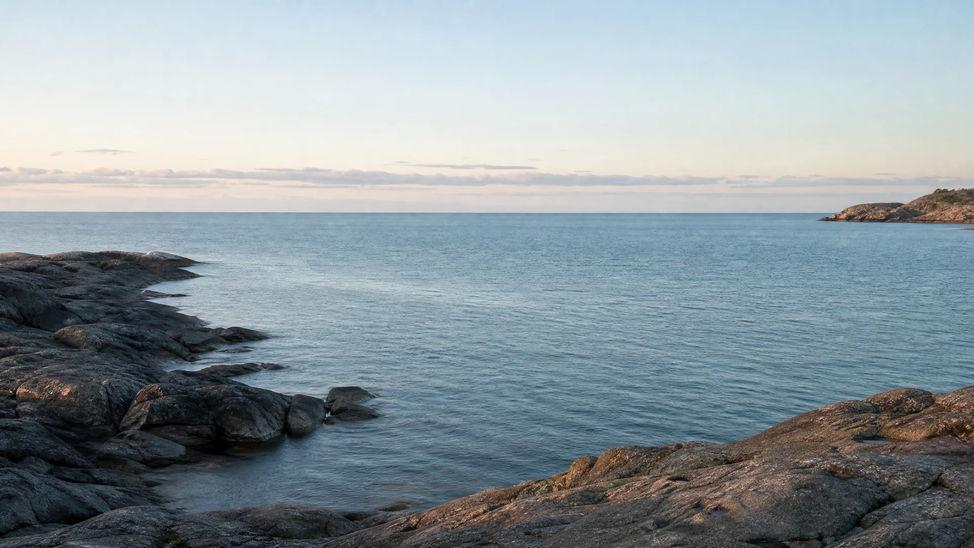 Calm sea under a pale clear sky with thin low clouds near the horizon, bordered by smooth bare rock outcrops in the foreground and a low rocky point in the distance at right.