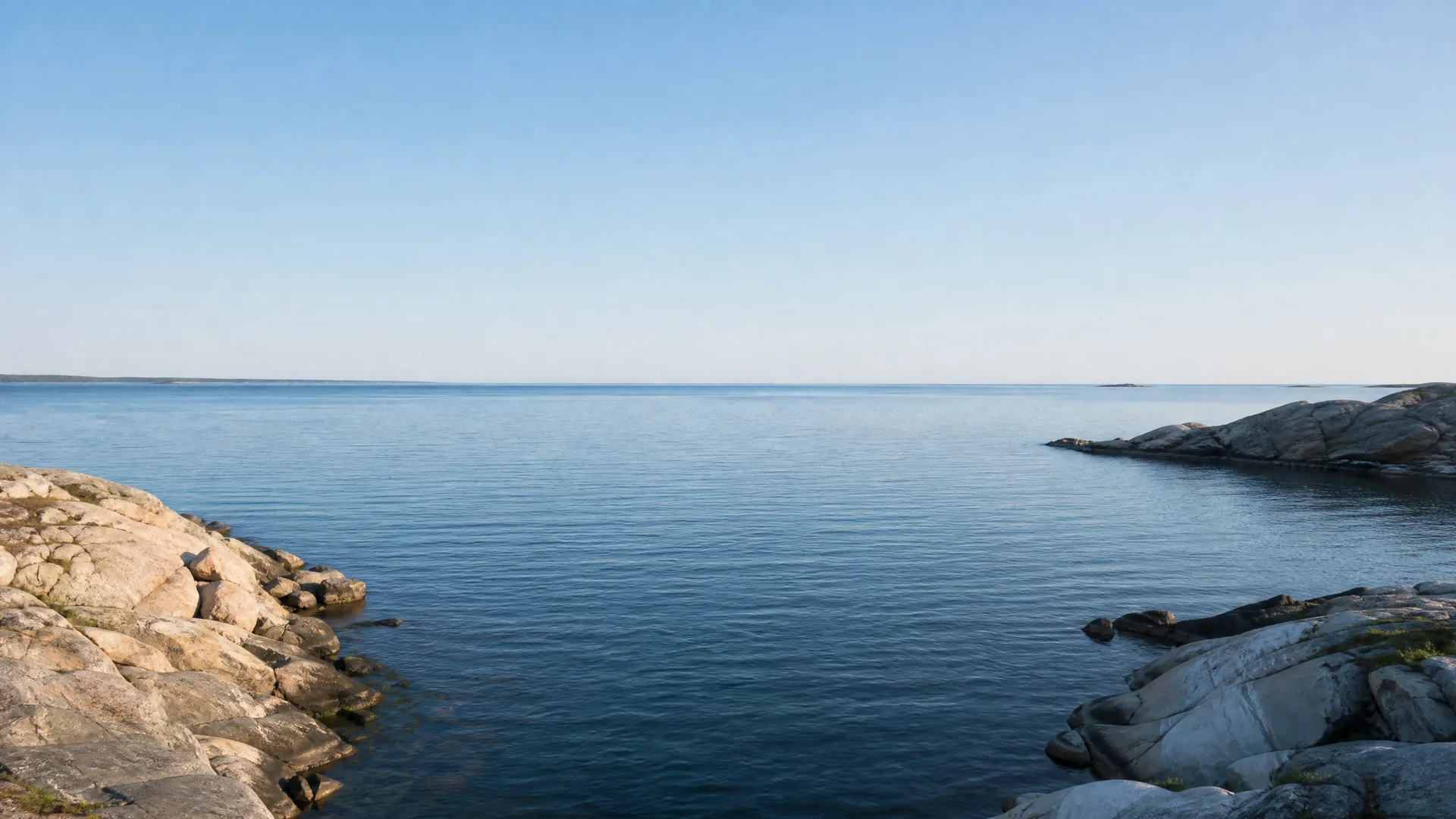 A calm sea under a clear blue sky, framed by low bare rocky shorelines in the left and right foreground with a distant flat coastline on the horizon.