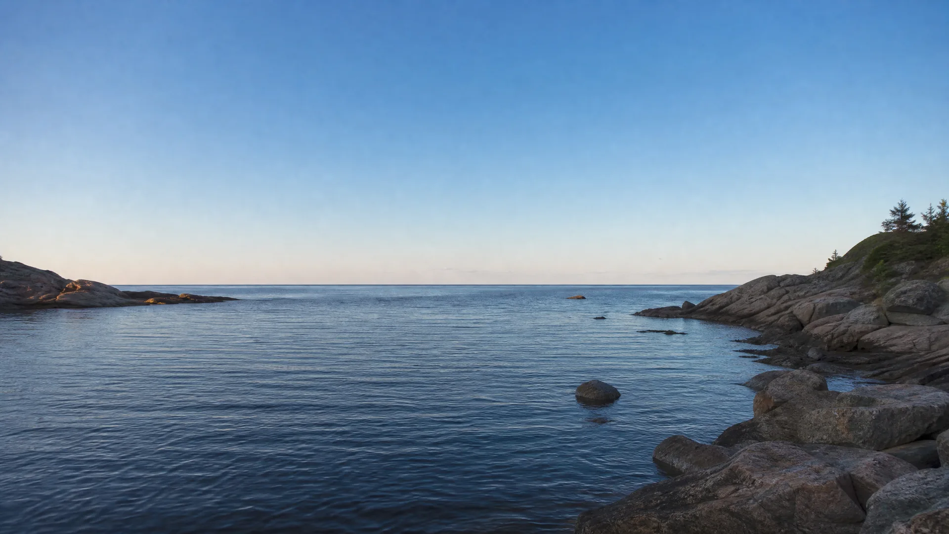 Calm water in a rocky cove under a clear blue sky, with granite boulders along the right shore, a low rocky point on the left, and a few conifers on the upper right.