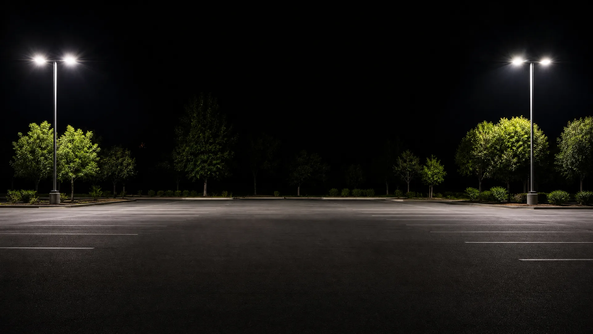 An empty asphalt parking lot at night, lit by two tall double-headed light poles with trees and shrubs along the far edge.