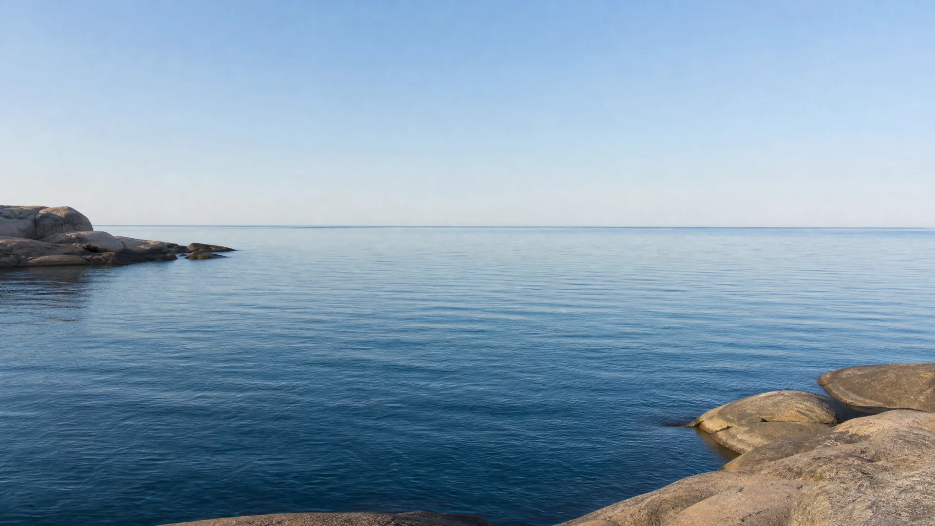 Calm blue water under a clear sky, with smooth bare rock outcrops in the lower right and left foreground and a flat horizon.