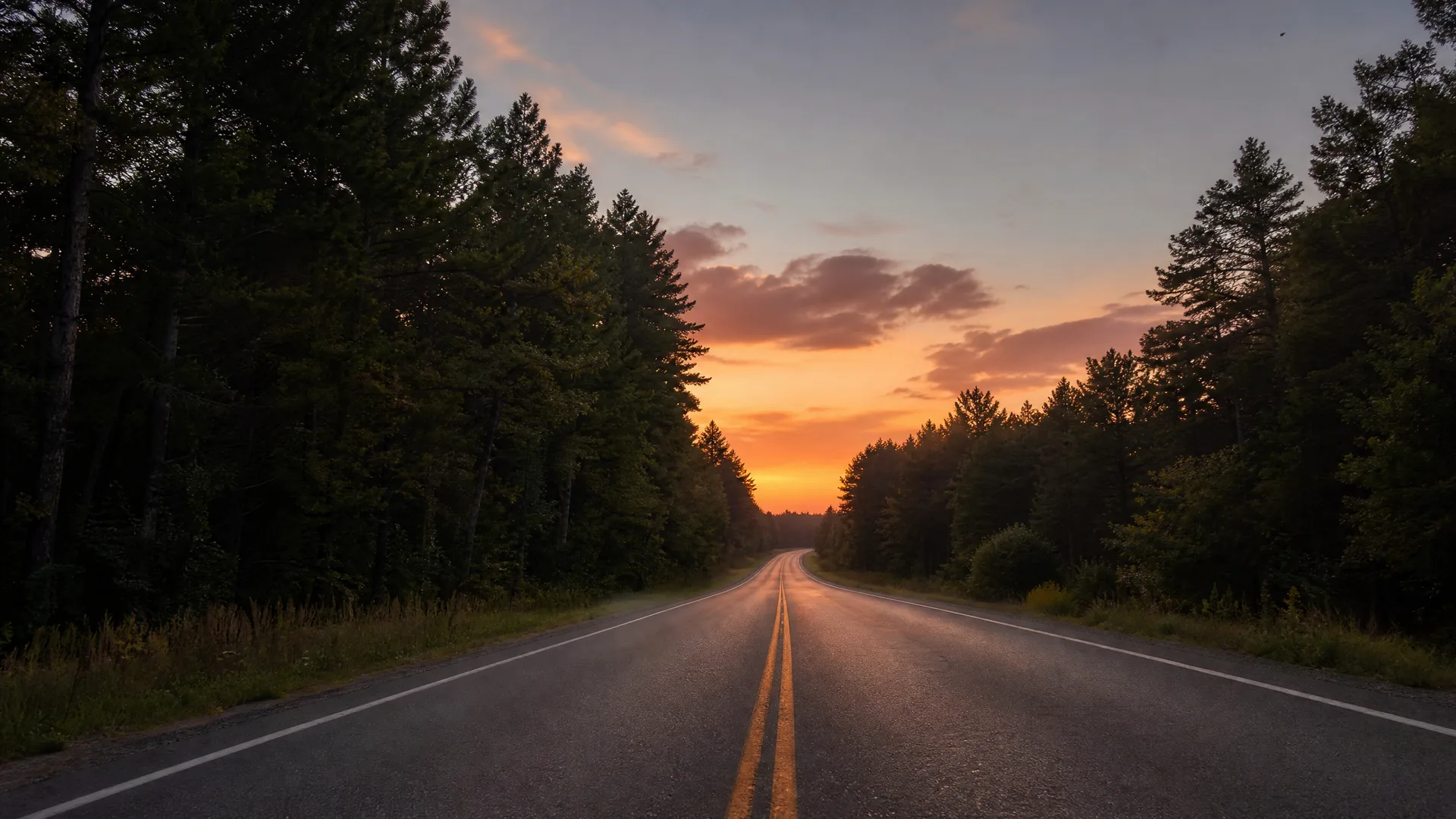 A two-lane asphalt road with double yellow center lines runs toward a distant bend between dark conifer forest, under an orange and purple sunset sky.
