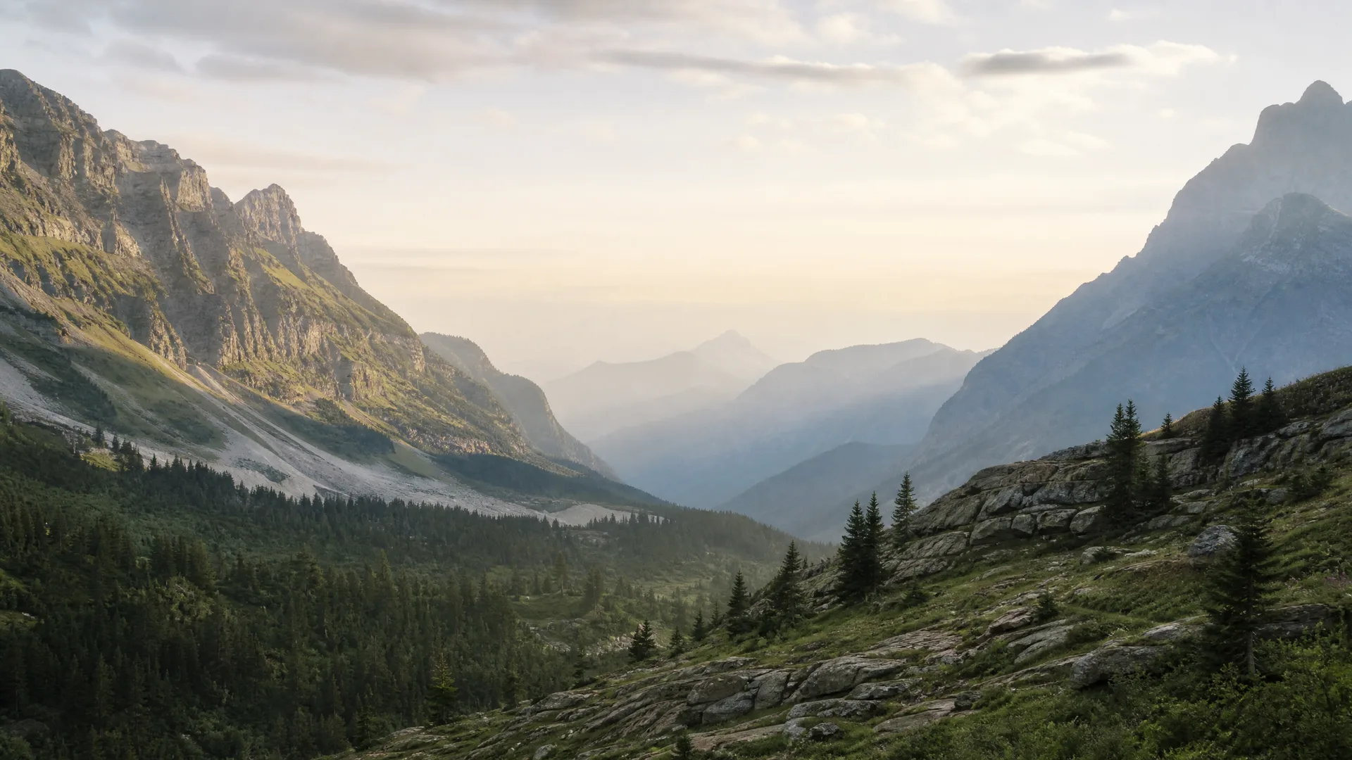 A hazy mountain valley at low sun, with rocky peaks on both sides, conifer forest and grassy rock slopes in the foreground, and receding ridgelines in the distance.