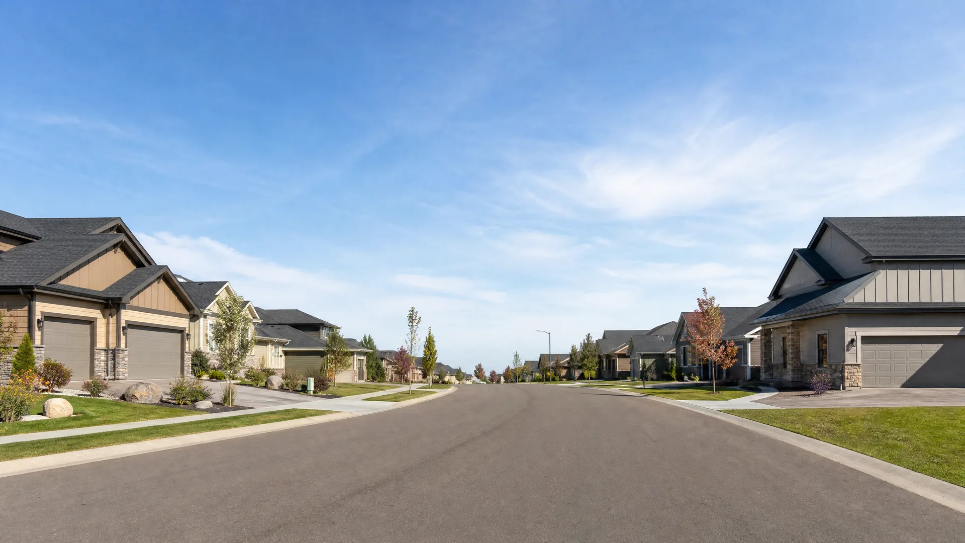 A wide empty asphalt street lined with single-family houses with attached garages, stone accents, young trees, and mown lawns under a mostly clear blue sky.