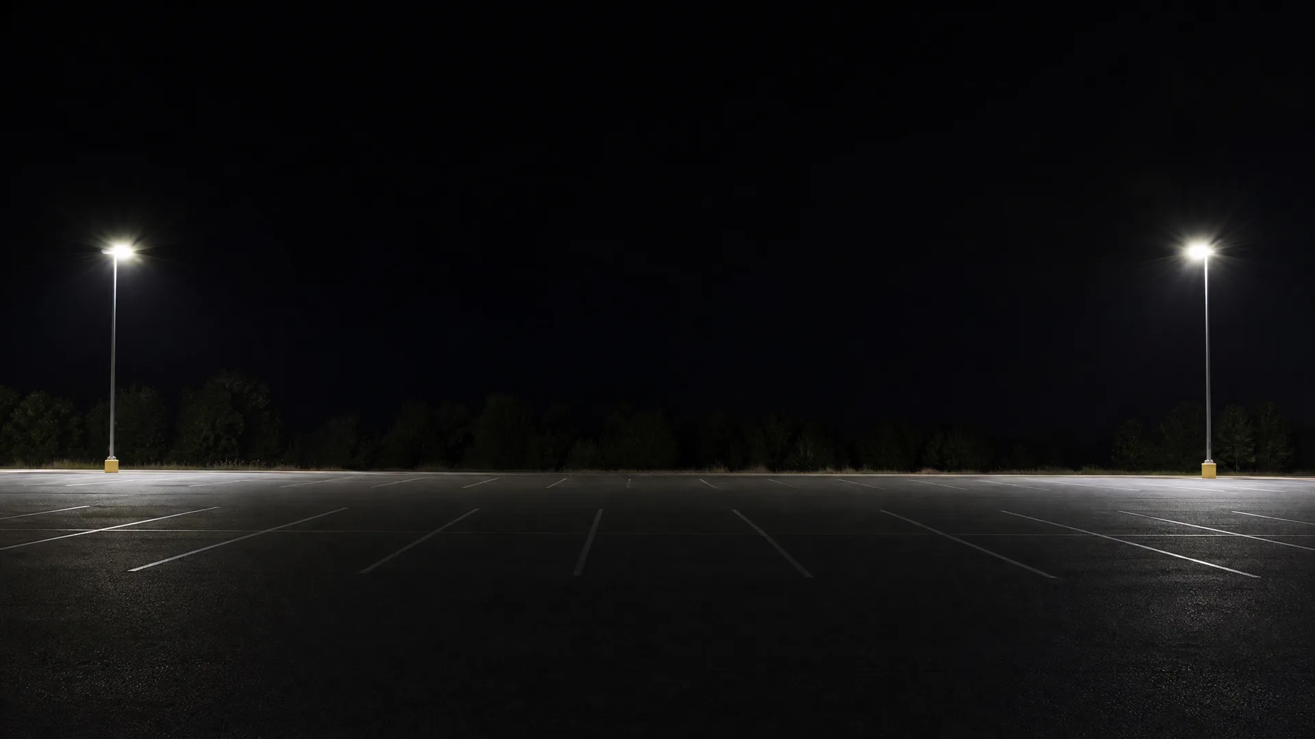 An empty asphalt parking lot at night with painted white stall lines, lit by two tall light poles on either side, with a dark tree line and black sky behind.