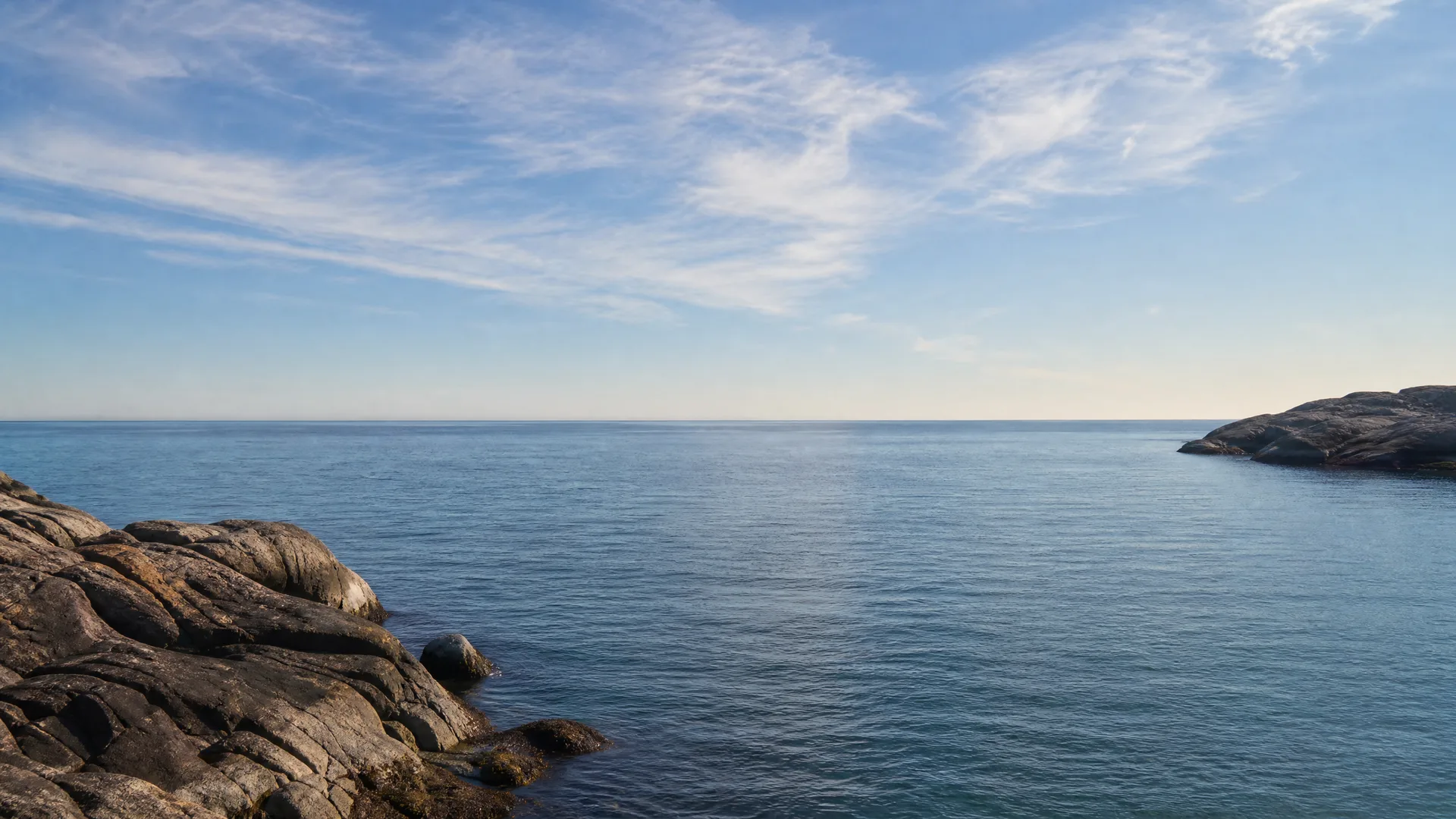 Calm blue sea under a clear sky with thin wispy clouds, bordered by bare rocky outcrops in the lower left foreground and a low rocky headland on the right.