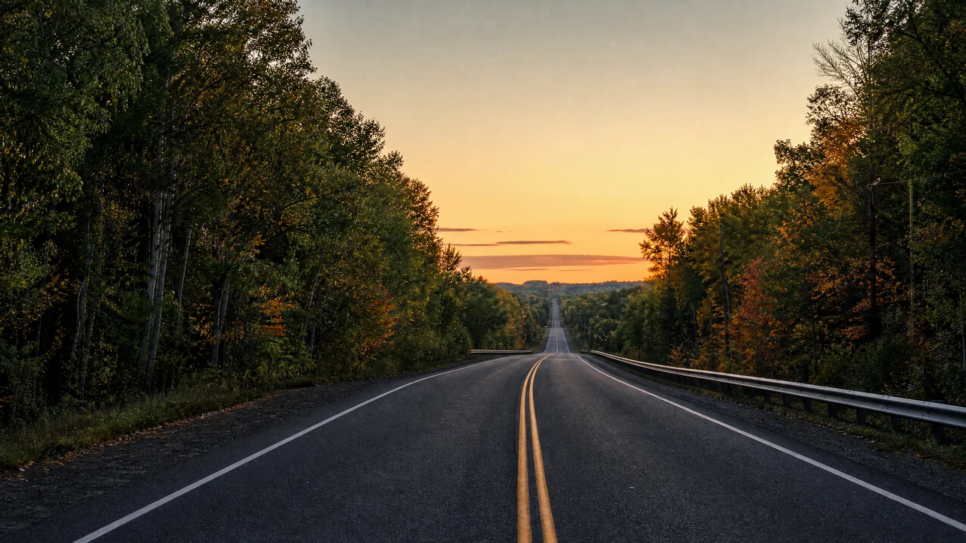An empty two-lane asphalt road with double yellow center lines runs straight toward a distant horizon between dense trees, under an orange and blue sky at dusk, with a metal guardrail on the right.