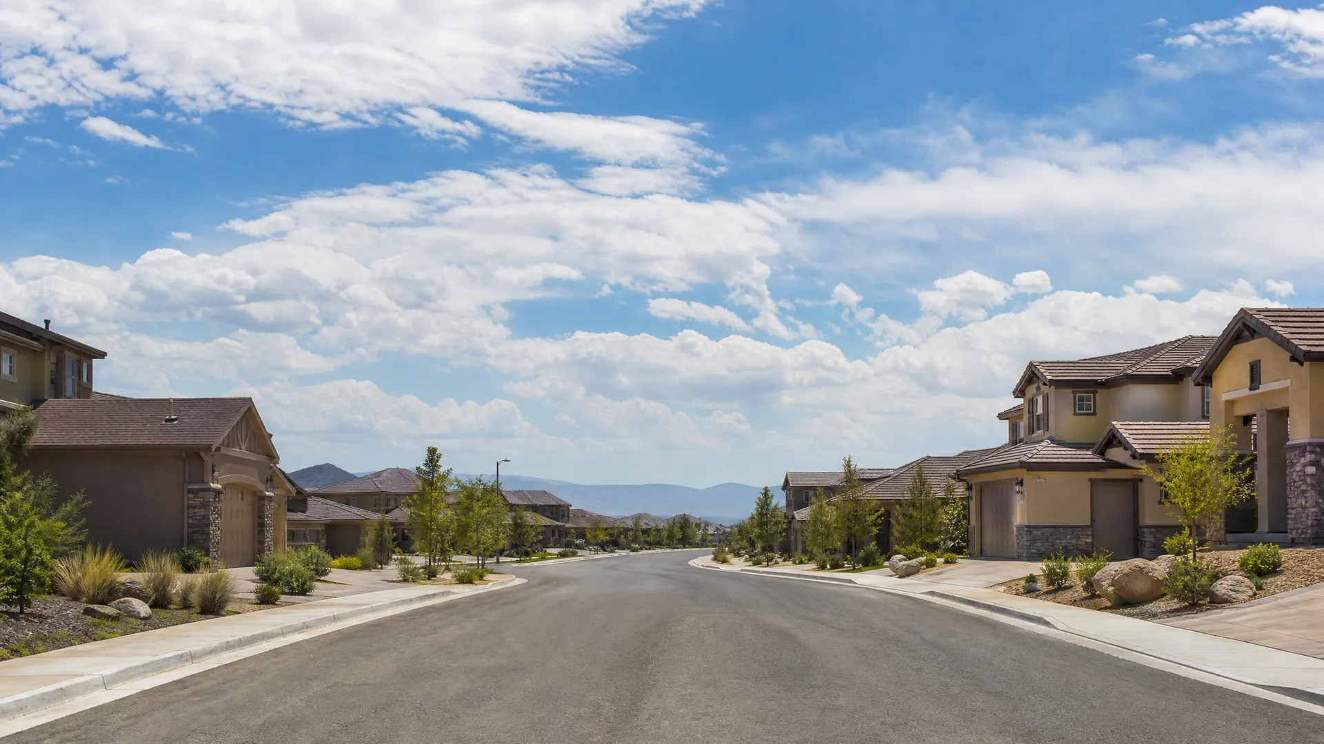 An empty paved residential street lined with stucco two-story houses, young trees, and landscaped rock beds, with distant mountains and a partly cloudy sky.