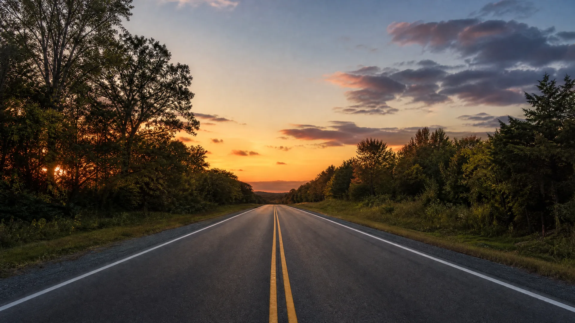 A two-lane asphalt road with double yellow center lines and white edge lines runs straight toward the horizon between tree-lined shoulders under an orange and blue sunset sky.