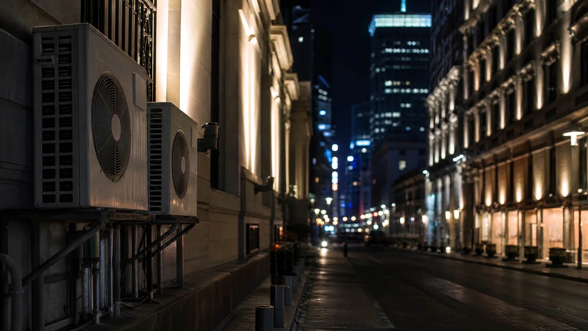 Night view down a city street lined with lit stone buildings, with two air conditioning condenser units mounted on a wall in the left foreground and blurred lights of taller buildings in the distance.