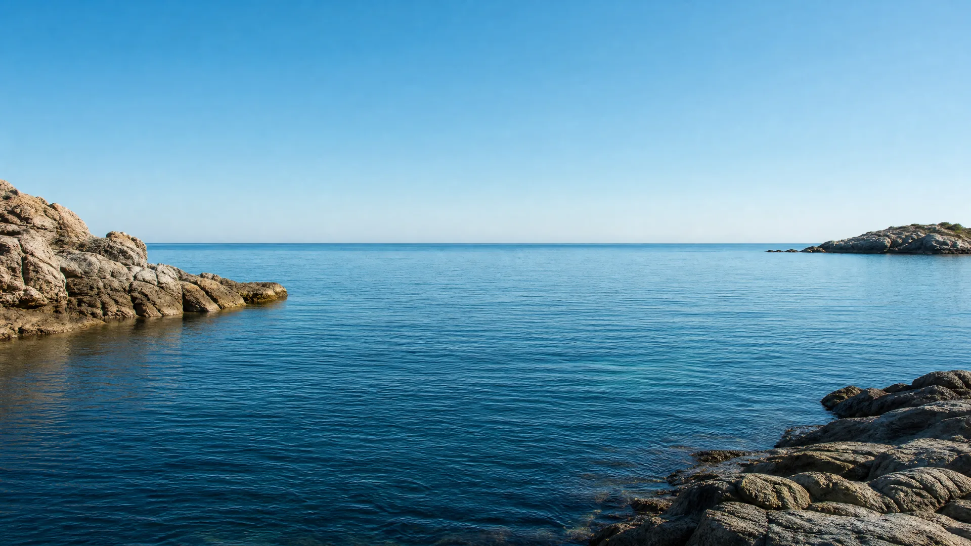 A calm blue sea under a clear sky, framed by rocky shorelines in the left foreground and lower right, with a low headland on the right horizon.