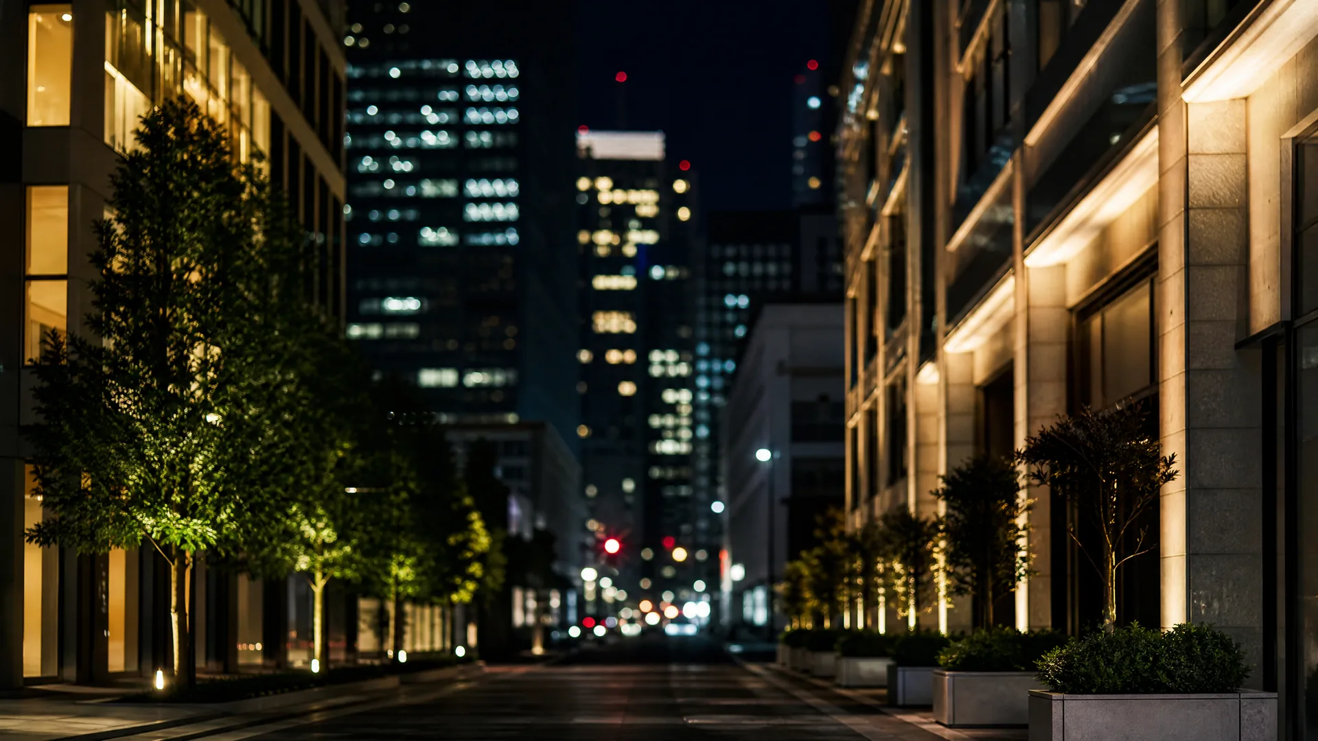 A nighttime city street lined with young trees and planters, flanked by illuminated office buildings, with blurred lit high-rises in the distance.