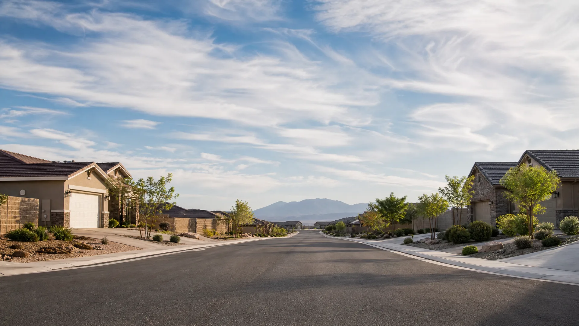 An empty paved residential street lined with single-story stucco houses, young trees and low shrubs, with a mountain ridge and cloud-streaked sky in the distance.