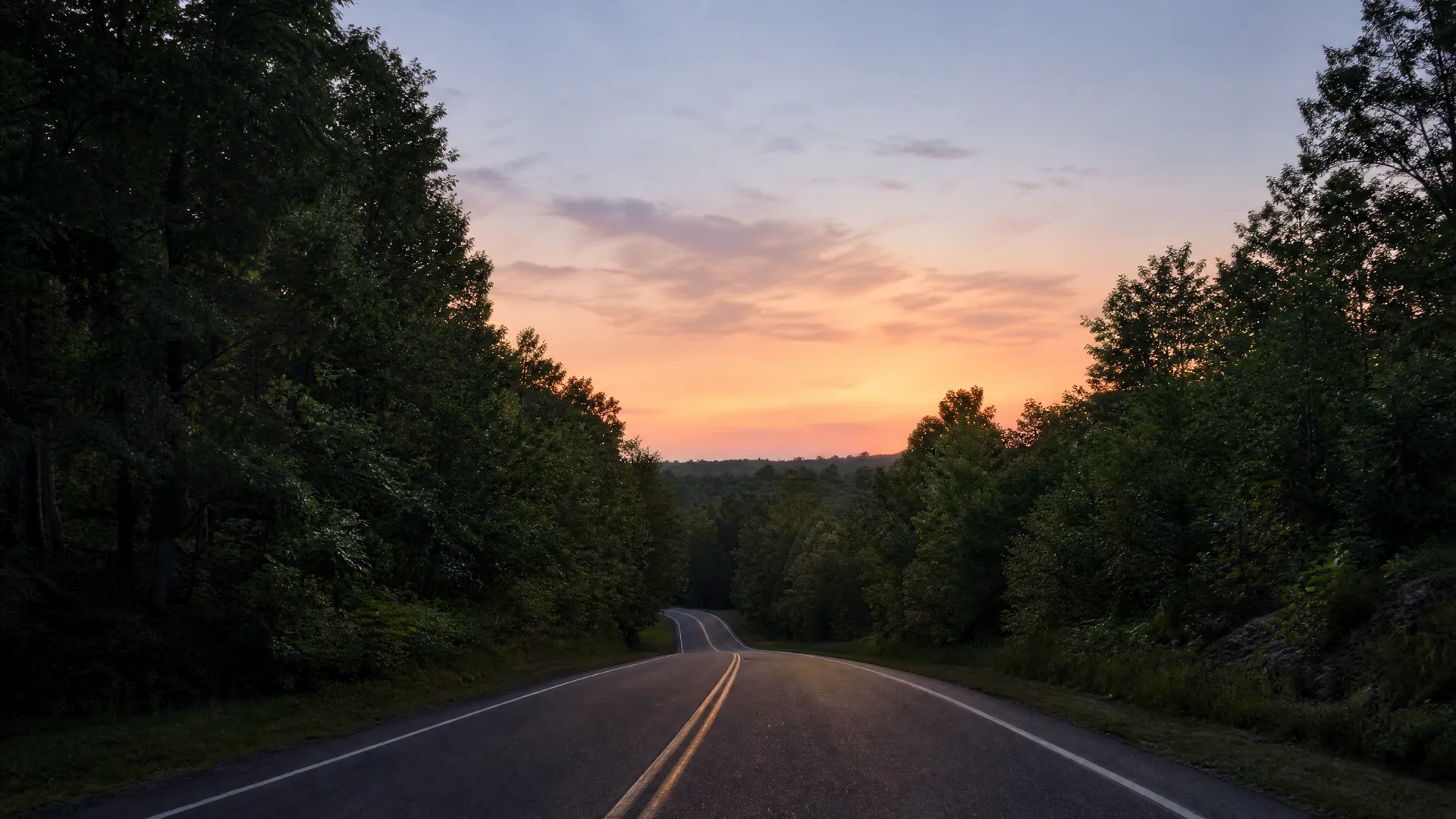 A two-lane paved road with double yellow center lines curving between dense deciduous trees toward a distant tree-covered ridge under an orange and pink sunset sky.