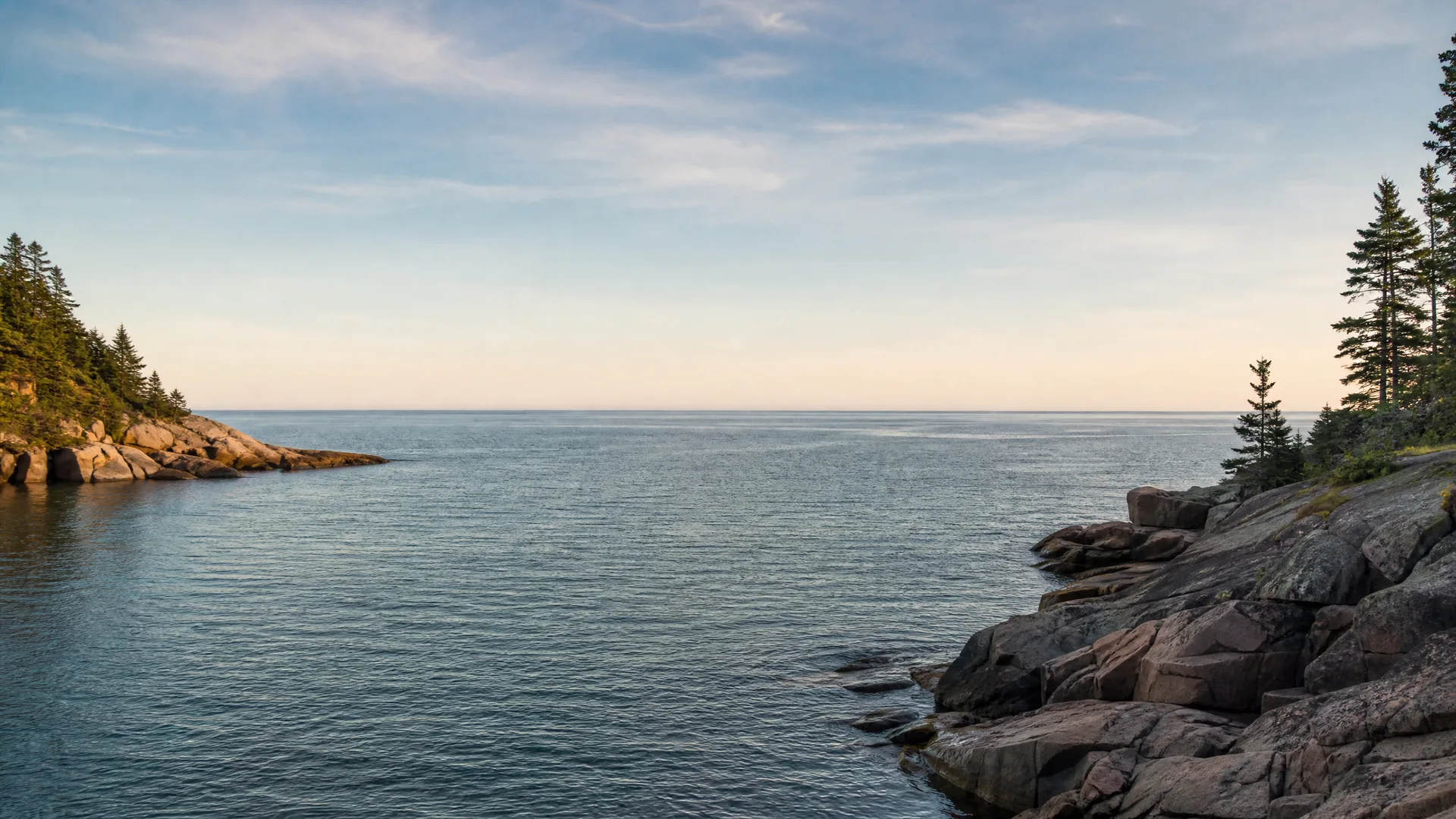 A calm bay seen from a rocky shoreline, with conifer trees on the headlands at left and right and a pale sky above a flat horizon.