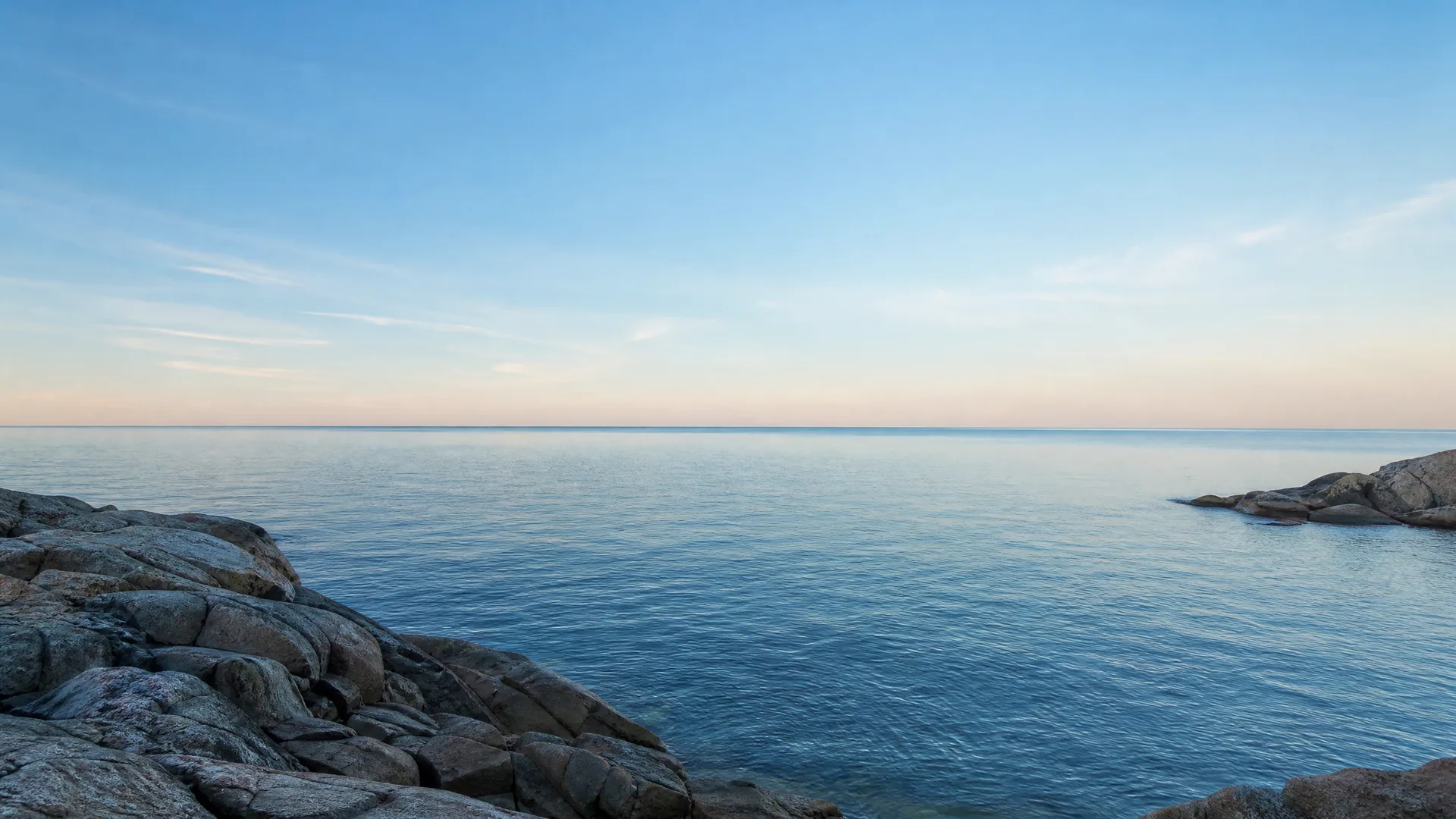 Calm blue sea under a pale clear sky at dusk, with bare rocky outcrops in the left foreground and along the right edge.