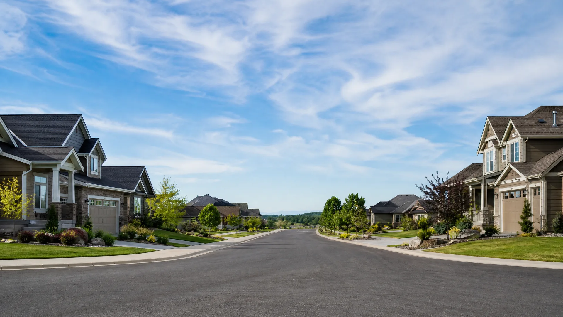 An empty paved residential street lined with two-story houses with attached garages, lawns, shrubs and young trees under a blue sky with thin clouds.