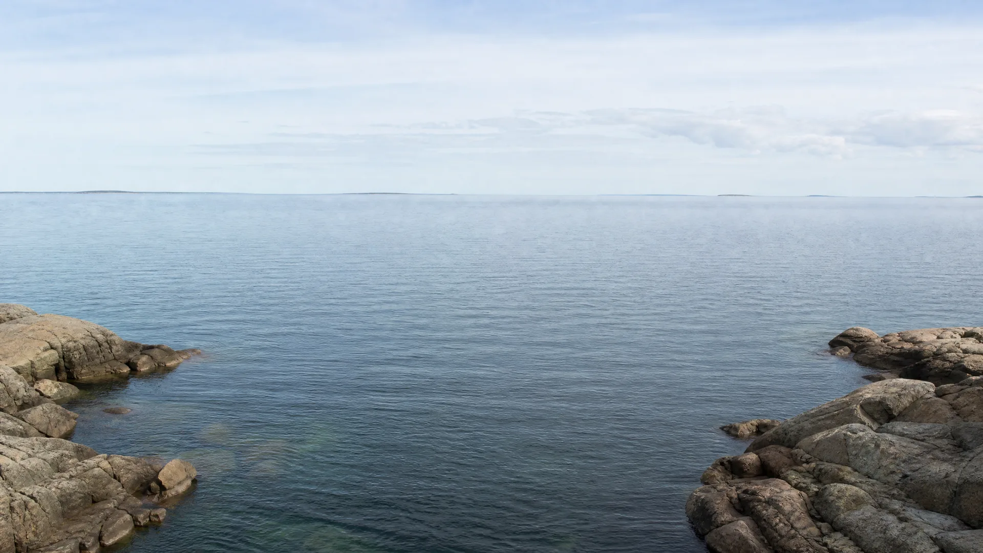 Calm open water seen between low rocky outcrops in the foreground, with a distant flat shoreline and hazy sky above.