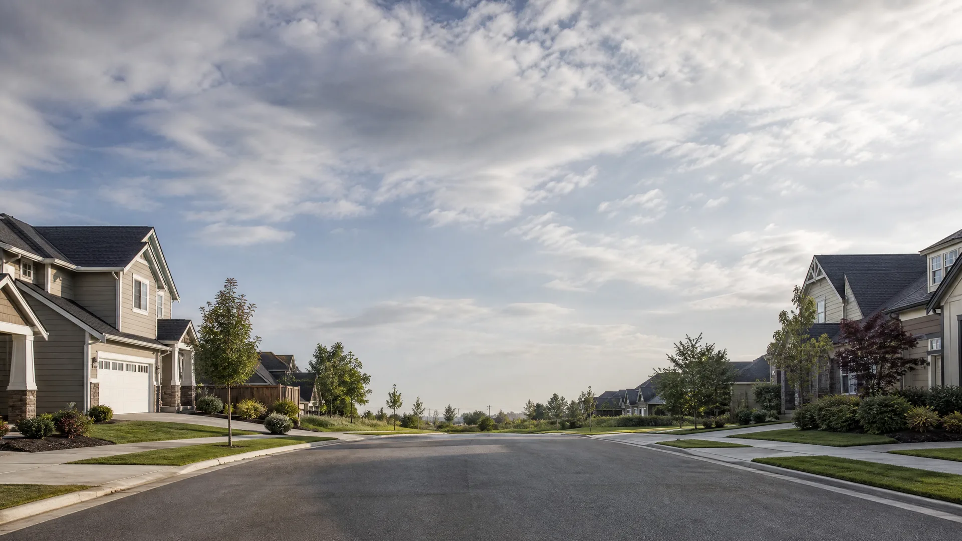 An empty paved residential street lined with two-story houses, sidewalks, lawns and young trees under a partly cloudy sky.