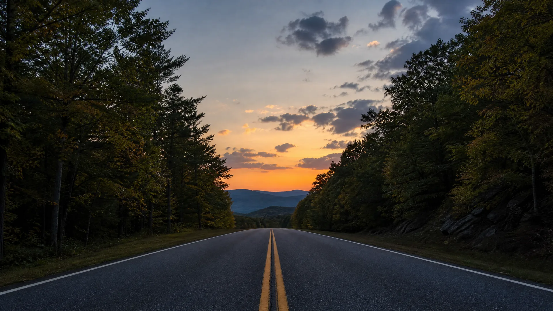 A two-lane asphalt road with a double yellow center line runs toward distant mountains, flanked by dark trees under a cloudy sky lit orange at sunset.