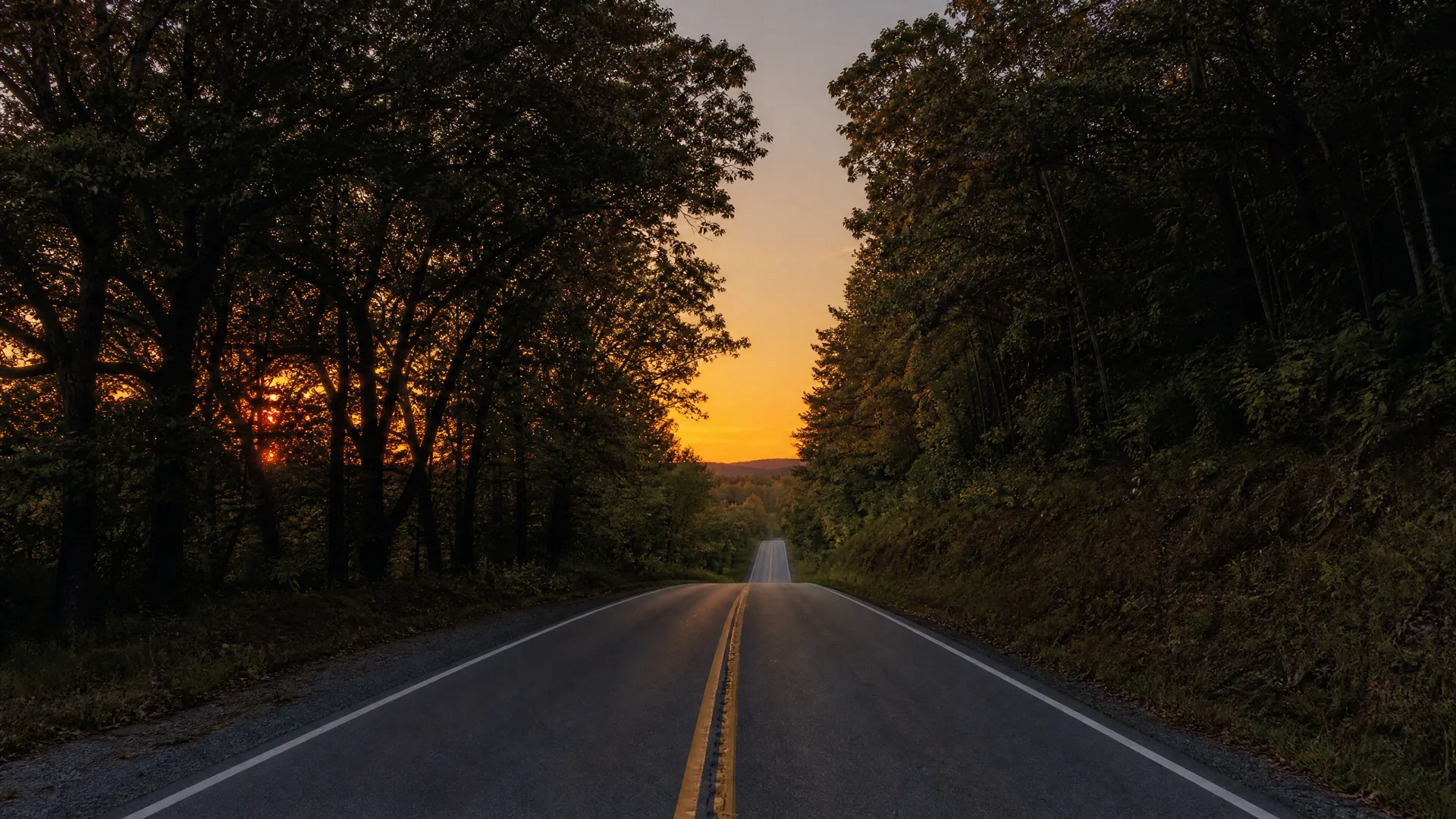 A two-lane paved road with a double yellow centerline runs straight toward a distant ridge, flanked by dark trees under an orange sunset sky.