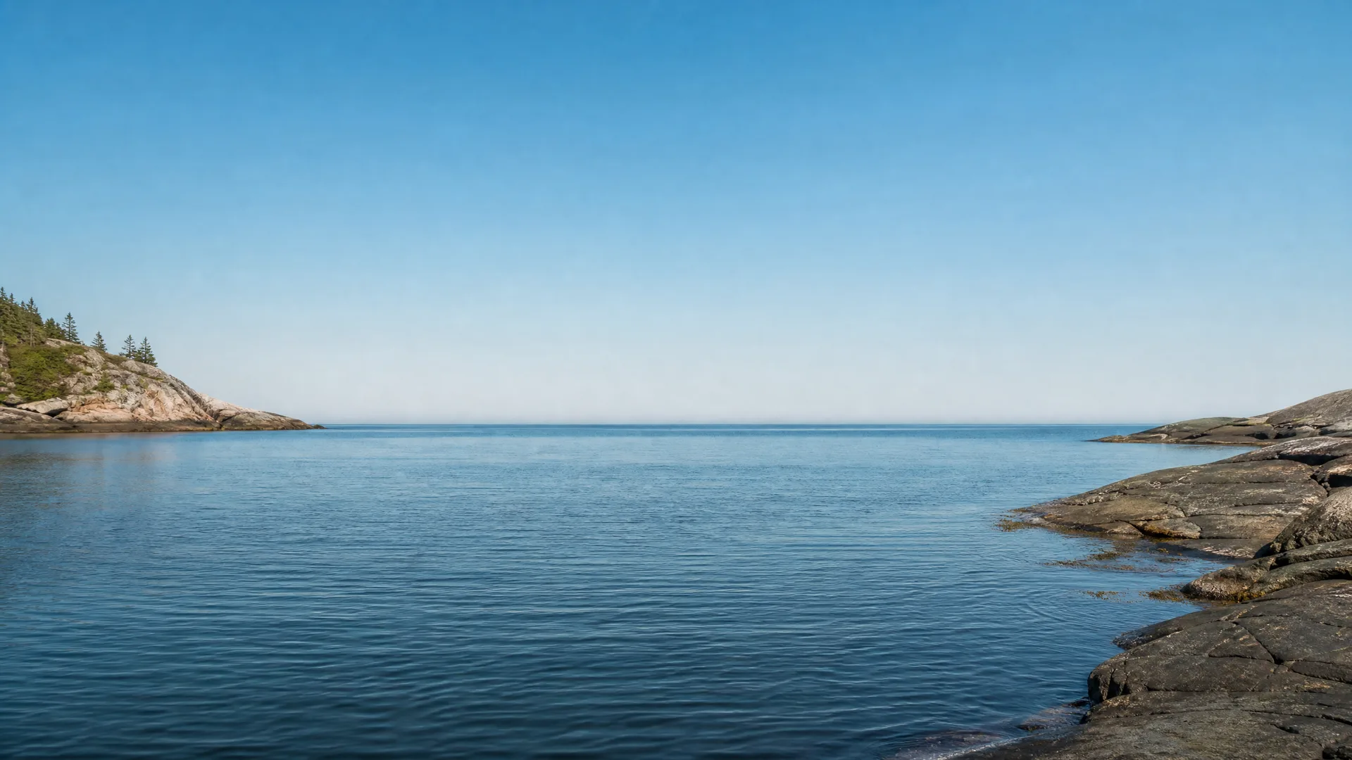 Calm blue water under a clear sky, framed by a rocky shoreline in the foreground right and a low wooded headland with conifers on the left.