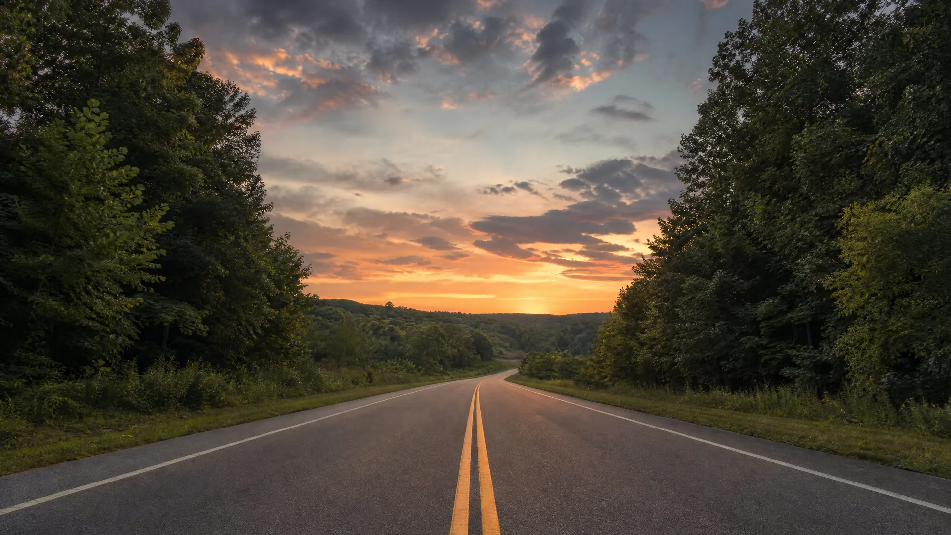 A two-lane asphalt road with double yellow center lines curves toward a tree-lined horizon under an orange and gray sunset sky.