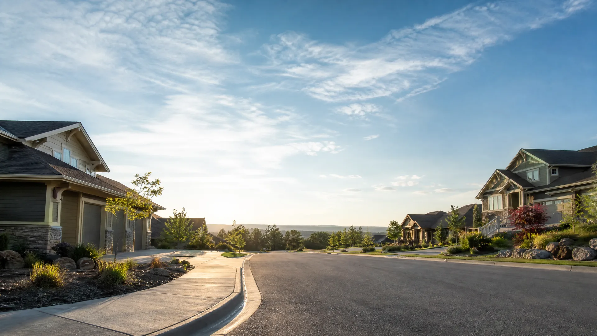 A paved suburban street curving between large single-family houses with stone and siding exteriors, landscaped yards and shrubs, with distant low hills and a partly cloudy sky at low sun.