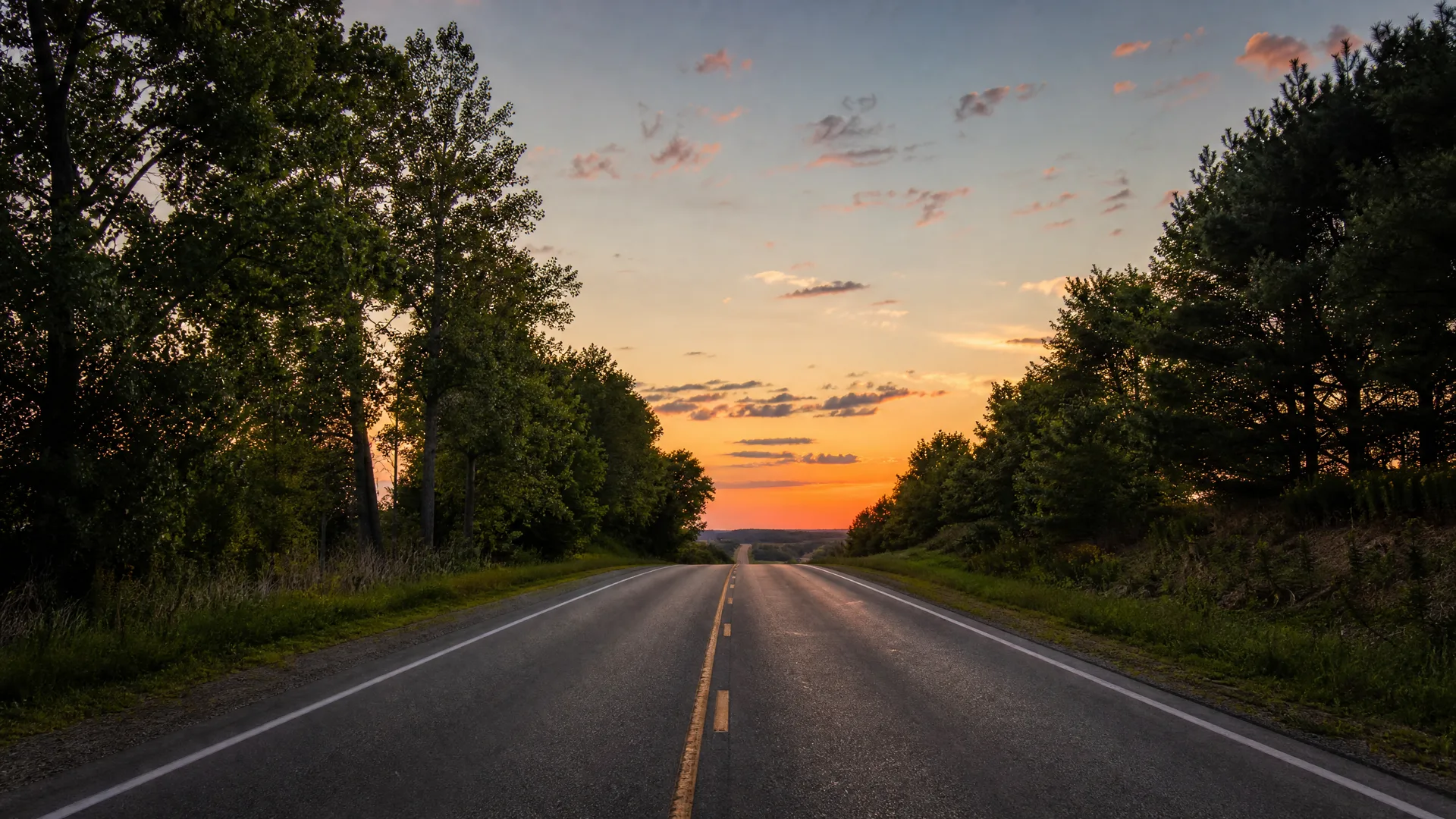 A two-lane asphalt road with a yellow center line runs toward the horizon between grassy verges and dense deciduous trees under an orange and blue sunset sky with scattered clouds.