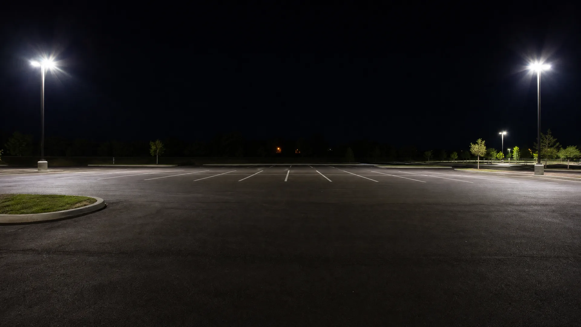An empty asphalt parking lot at night with painted white stall lines, lit by two tall pole-mounted lights and bordered by a grass curb and distant trees.