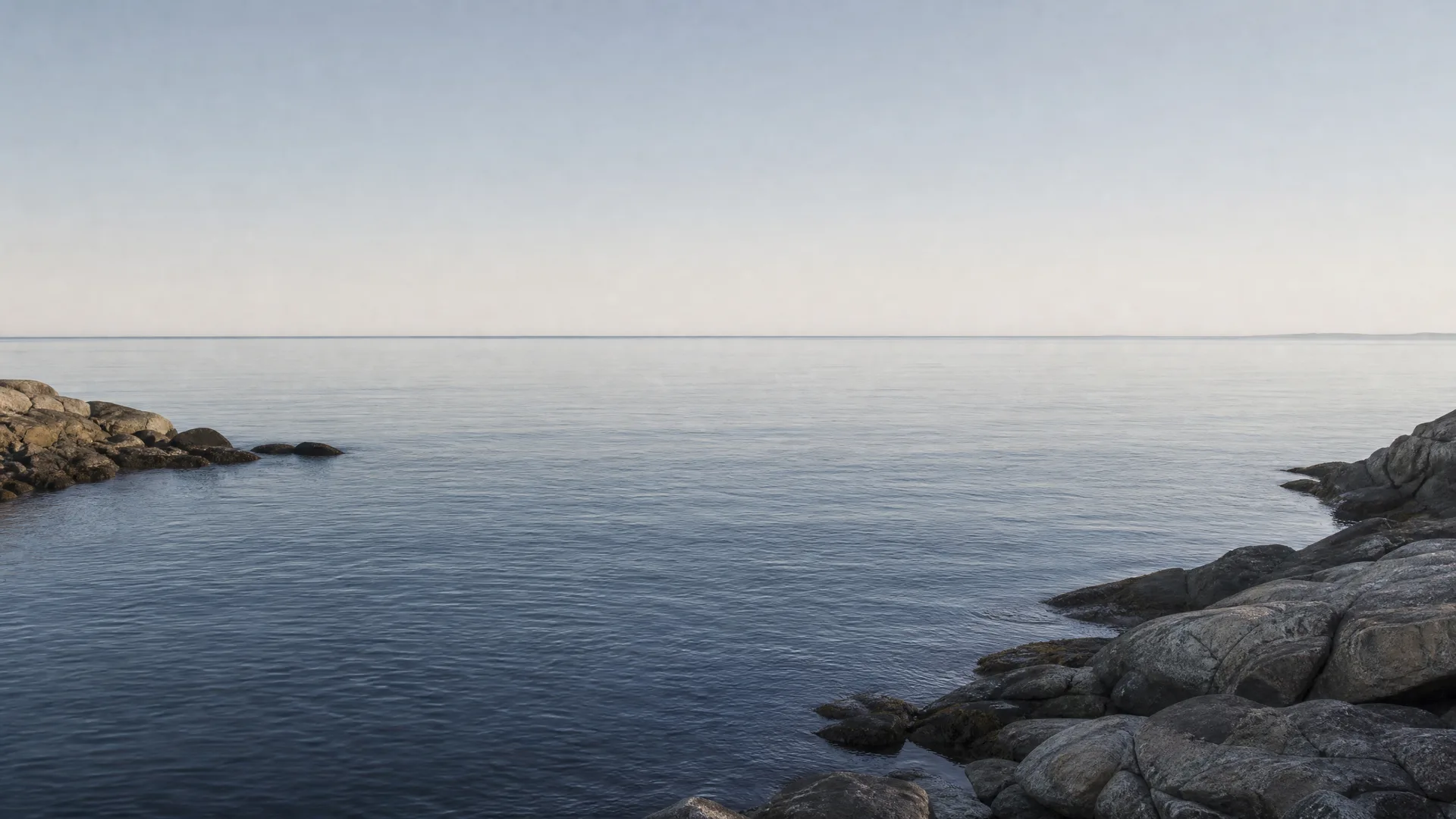 Calm water in a rocky cove under a pale clear sky, with low granite outcrops along the right and left edges and a faint distant shoreline on the horizon.