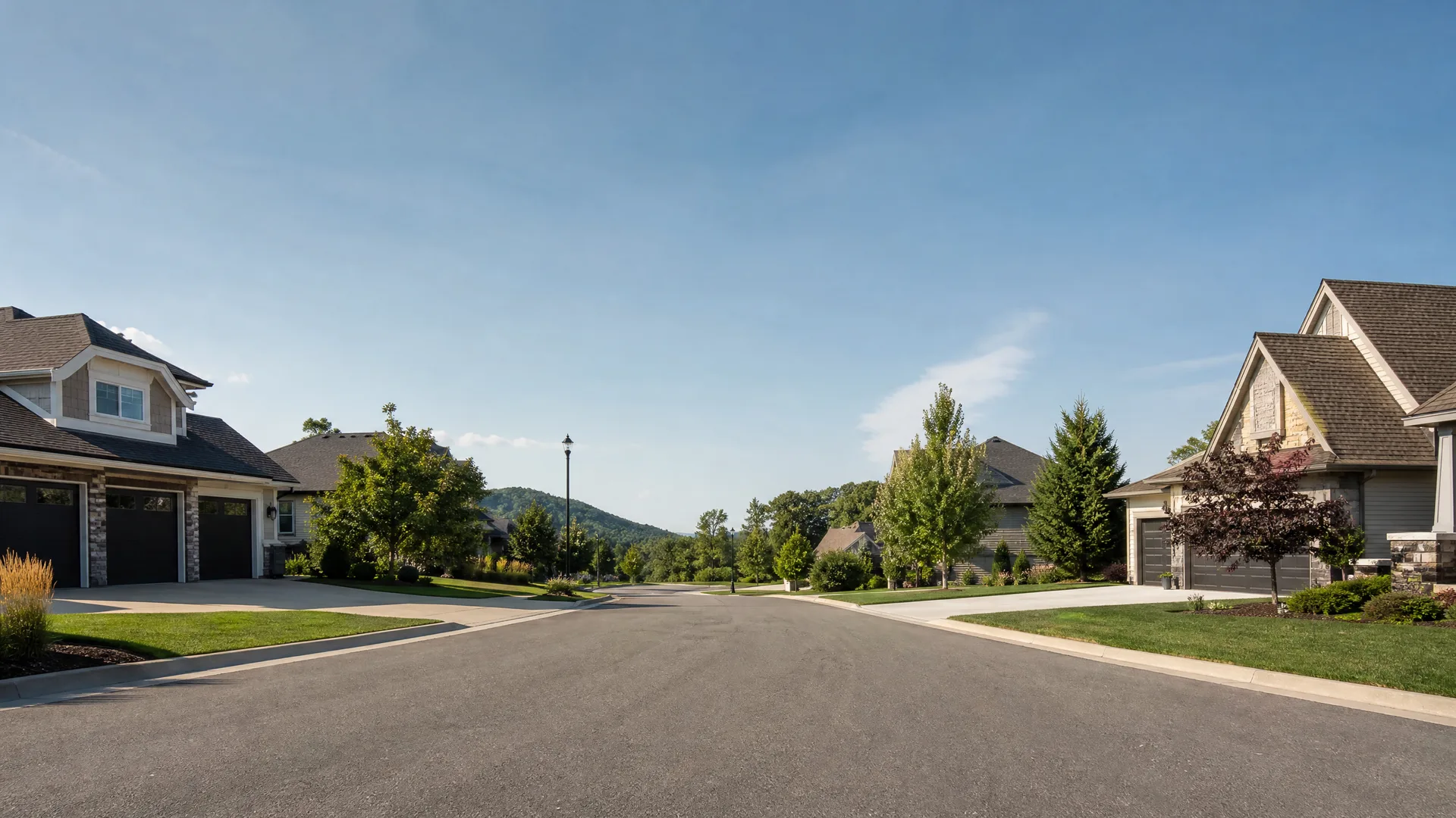A wide empty asphalt residential street lined with large houses with attached garages, lawns and young trees, with a low hill and clear blue sky in the distance.