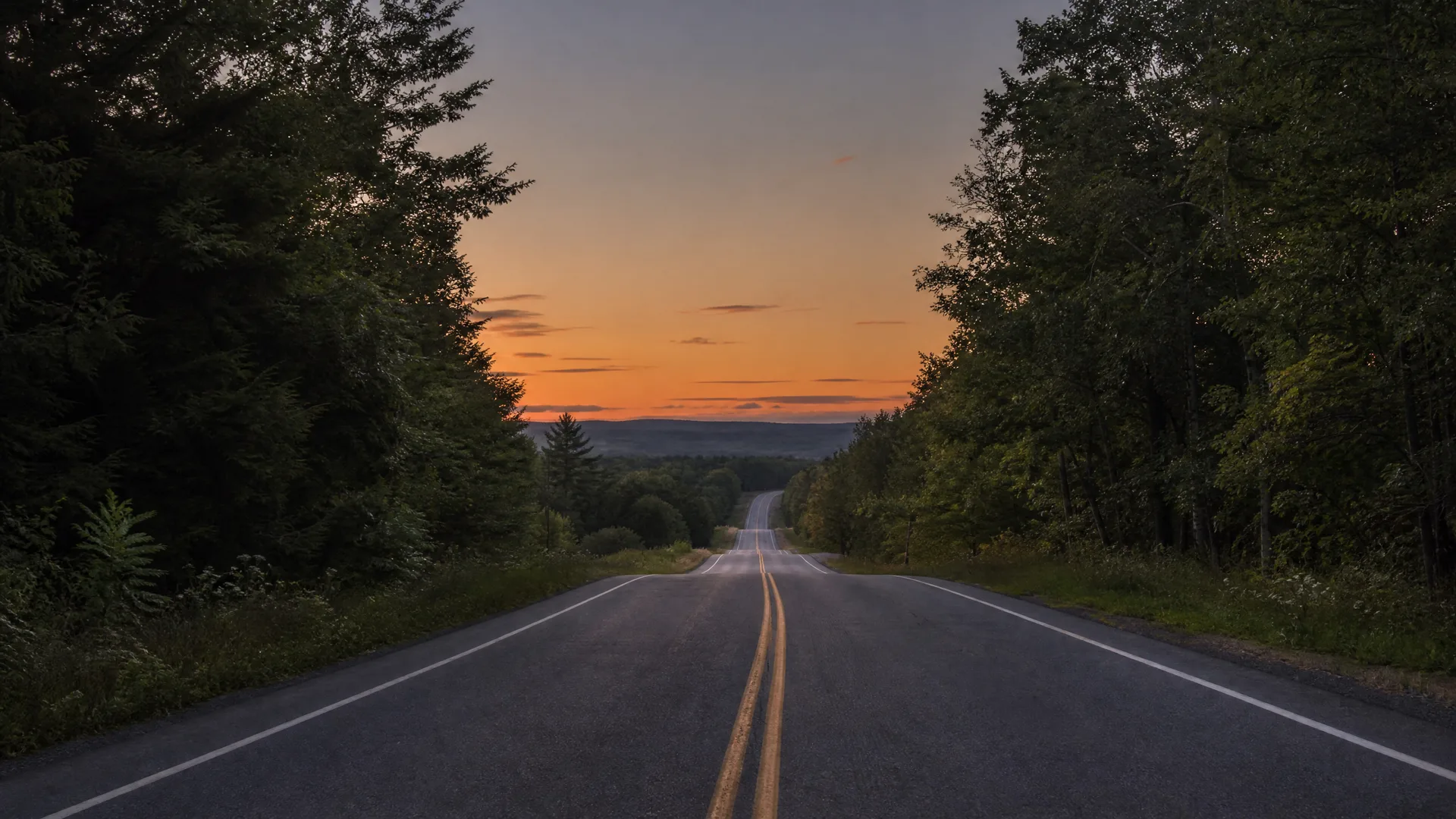 A two-lane road with a double yellow centerline and white edge lines runs straight toward low hills, bordered by dense trees under an orange sunset sky.