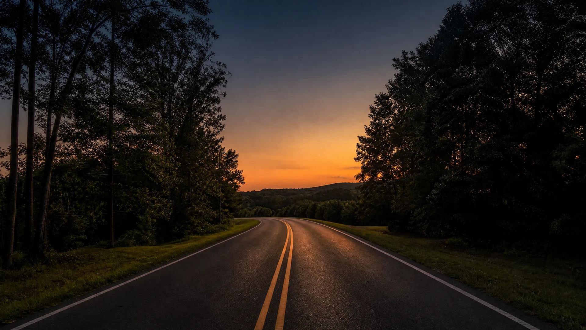 A two-lane asphalt road with double yellow center lines curves toward a distant tree-covered ridge under an orange and blue sunset sky, flanked by dark silhouetted trees.