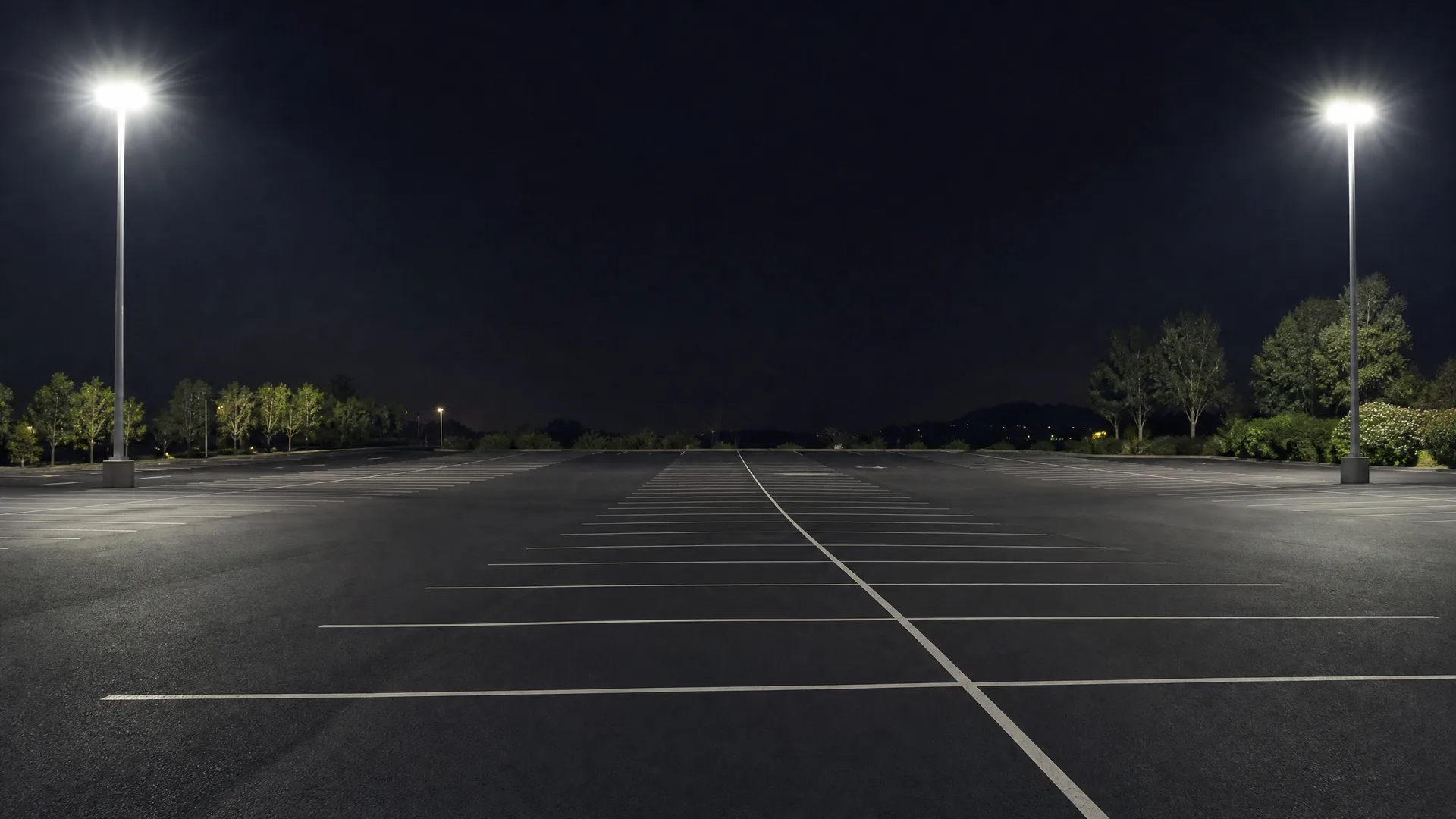 An empty asphalt parking lot at night with painted white stall lines, lit by two tall pole-mounted lights, bordered by trees under a dark sky.