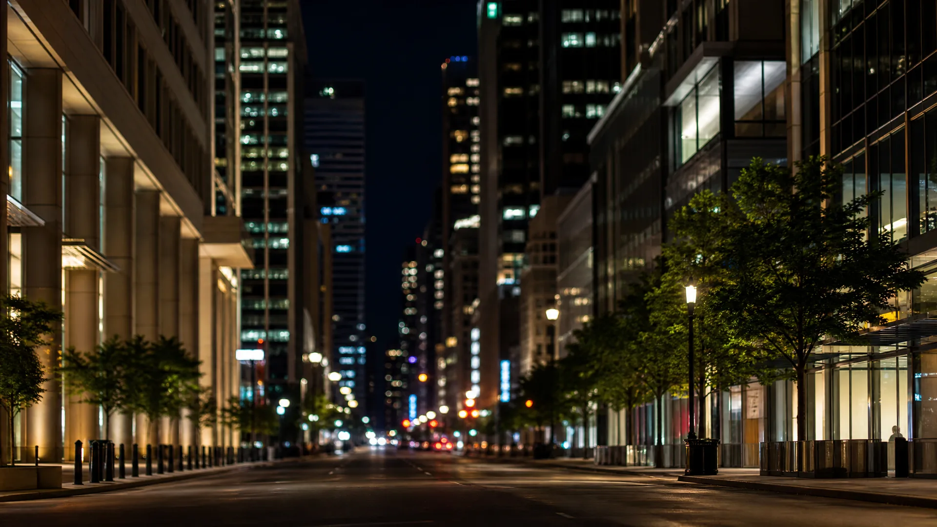 A wide empty city street at night lined with illuminated high-rise office buildings, street trees and a lamppost, with distant blurred lights down the corridor.