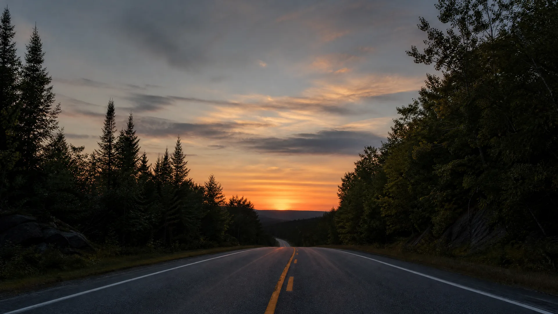 A two-lane paved road with a double yellow centre line runs toward low hills at sunset, bordered by dark conifer and deciduous trees under an orange and grey cloudy sky.