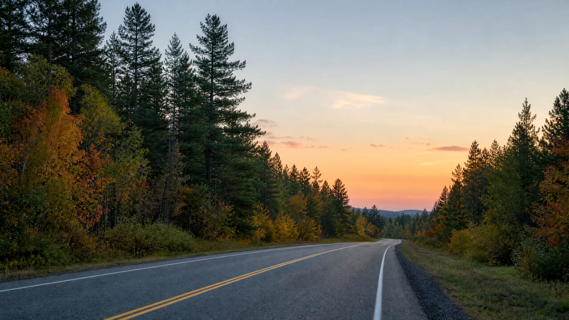 A two-lane paved road with a double yellow center line and white edge lines curves through conifer and deciduous forest with orange autumn foliage, under an orange and blue sky at dusk.