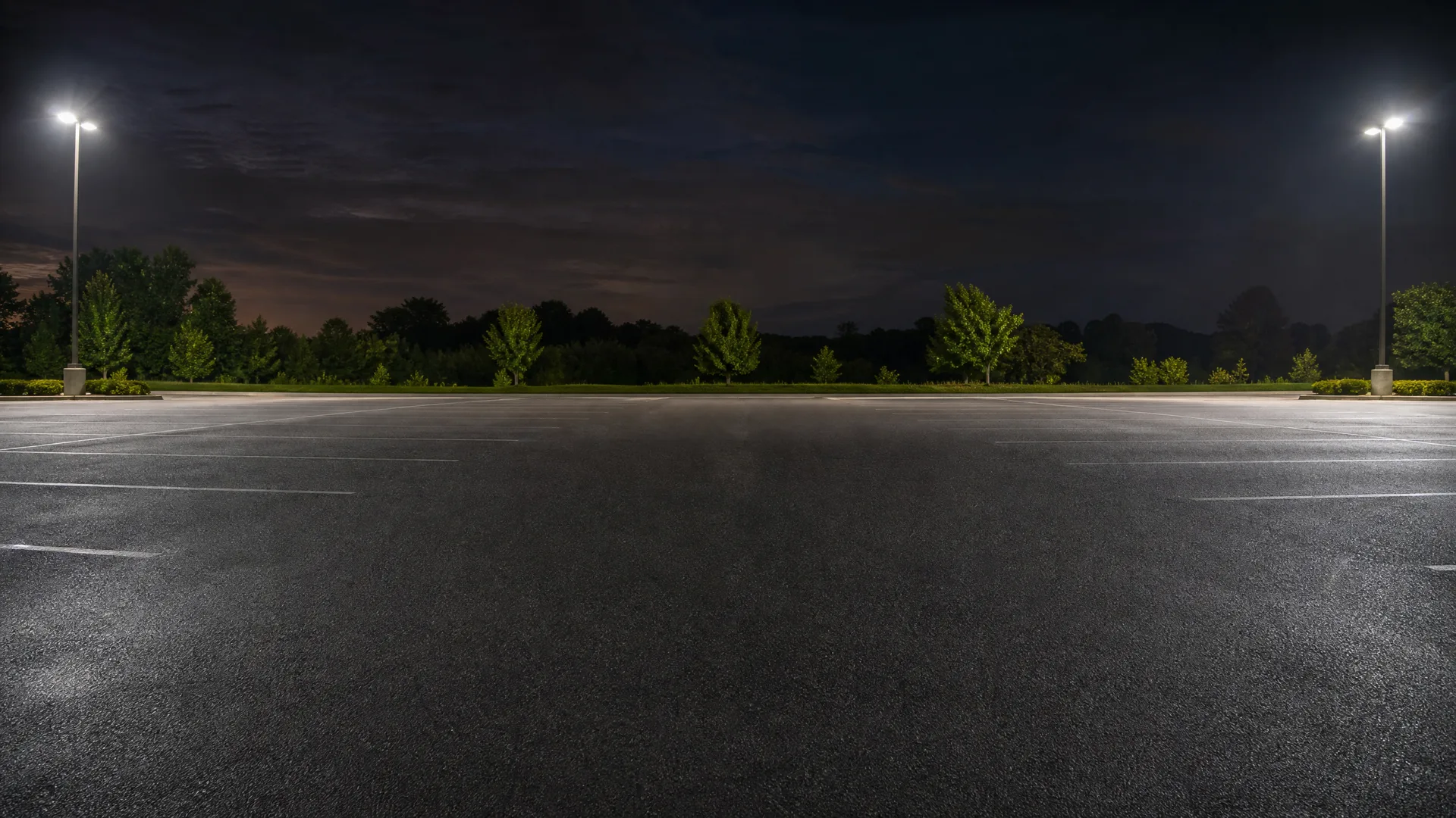 An empty asphalt parking lot at night with painted white stall lines, lit by two tall pole-mounted lights at left and right, with a treeline and dark sky behind.