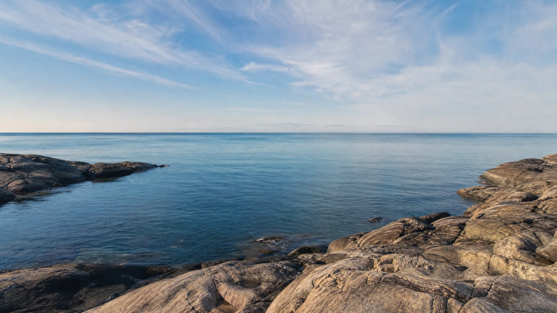 Smooth sloping rock shelves in the foreground and left frame the calm blue water of a bay under a pale sky with thin wispy clouds.