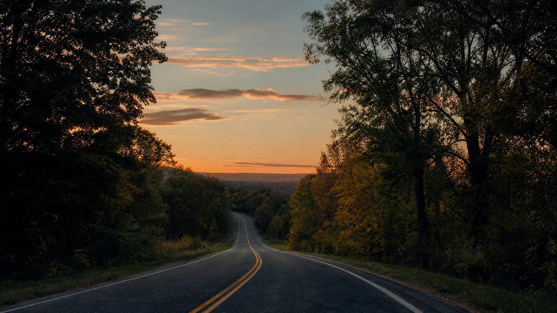 A two-lane paved road with a double yellow center line curves downhill between dense trees toward a distant valley under an orange sunset sky.