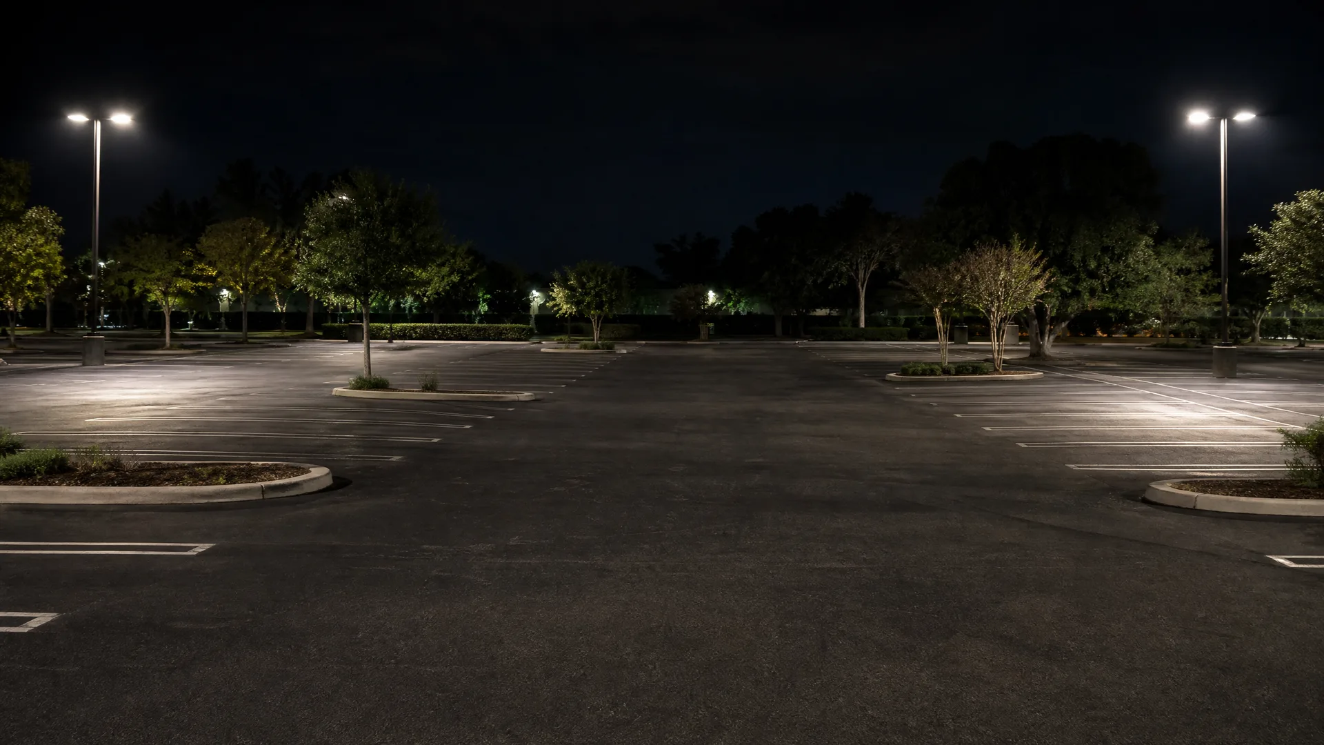 An empty asphalt parking lot at night, lit by two tall pole lights, with painted stall lines, curbed landscape islands and trees along the far edge.