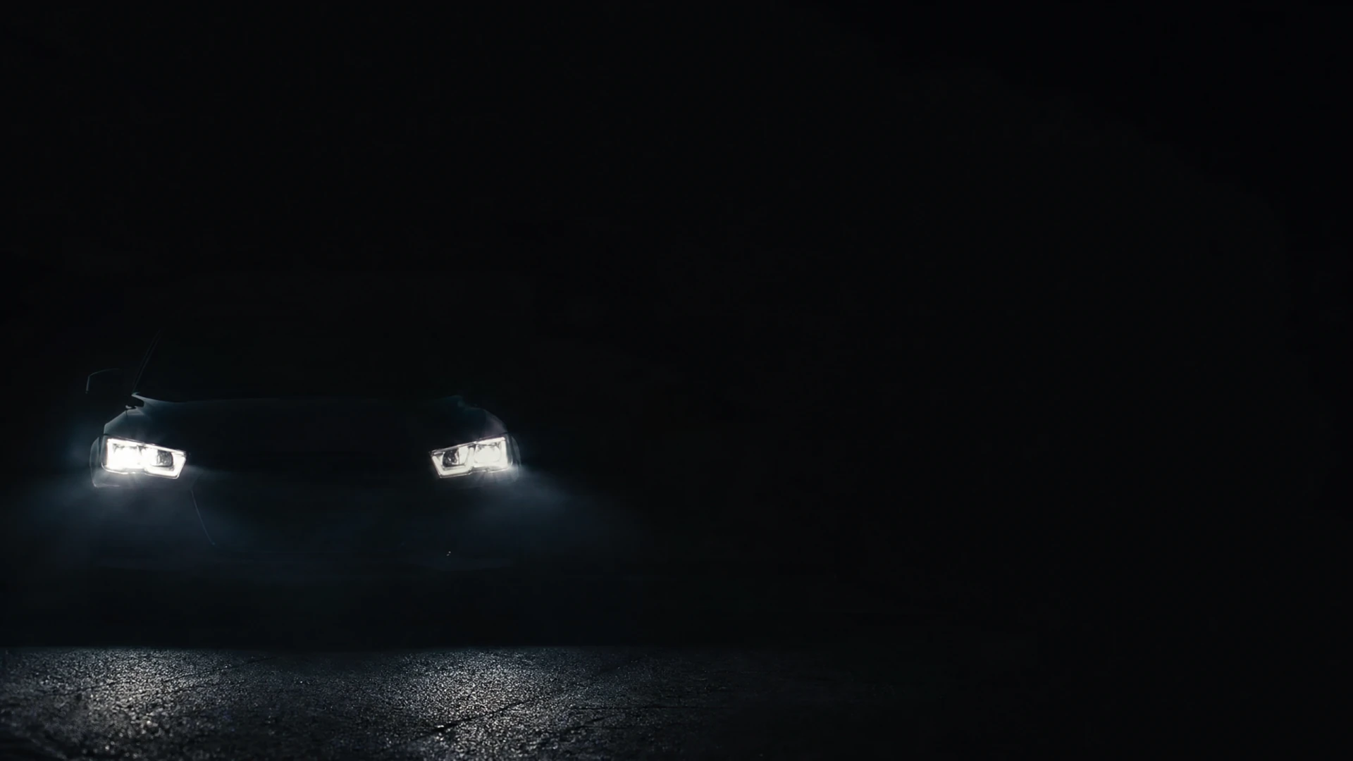 A dark car with two bright white headlights glowing in near-total darkness, casting light on wet pavement.
