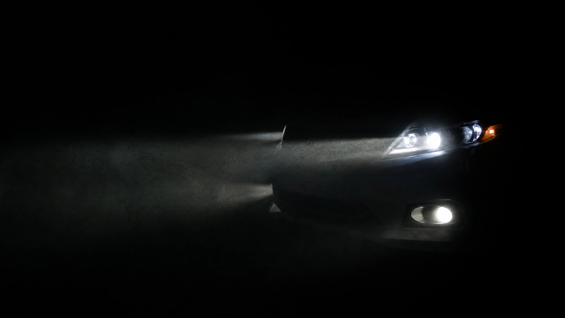 The front corner of a dark-colored car with glowing headlights and a fog light casting beams through hazy air against a black background.
