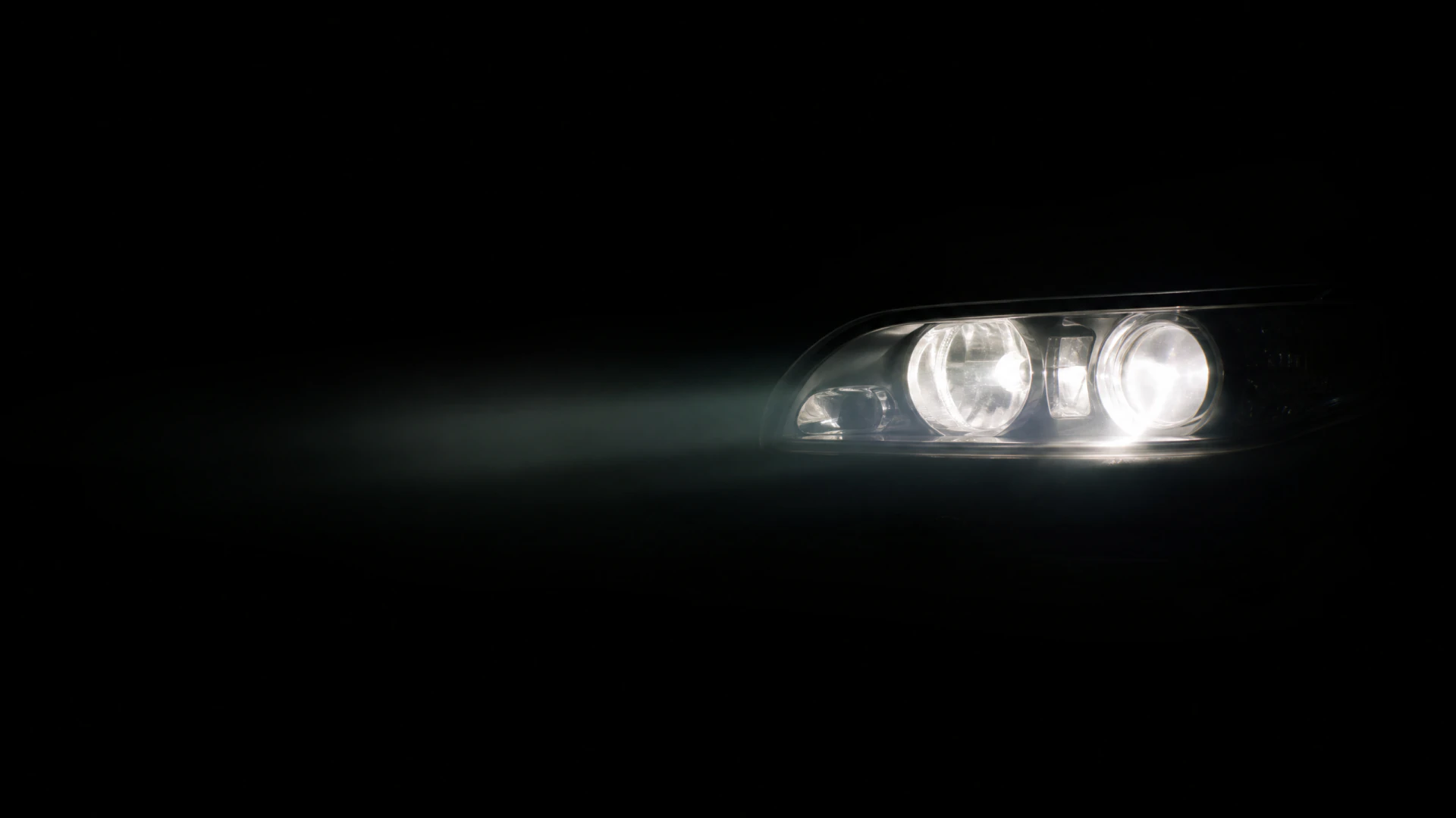 A close-up of a car's headlight assembly with two glowing circular projector bulbs casting a bright white beam into darkness.