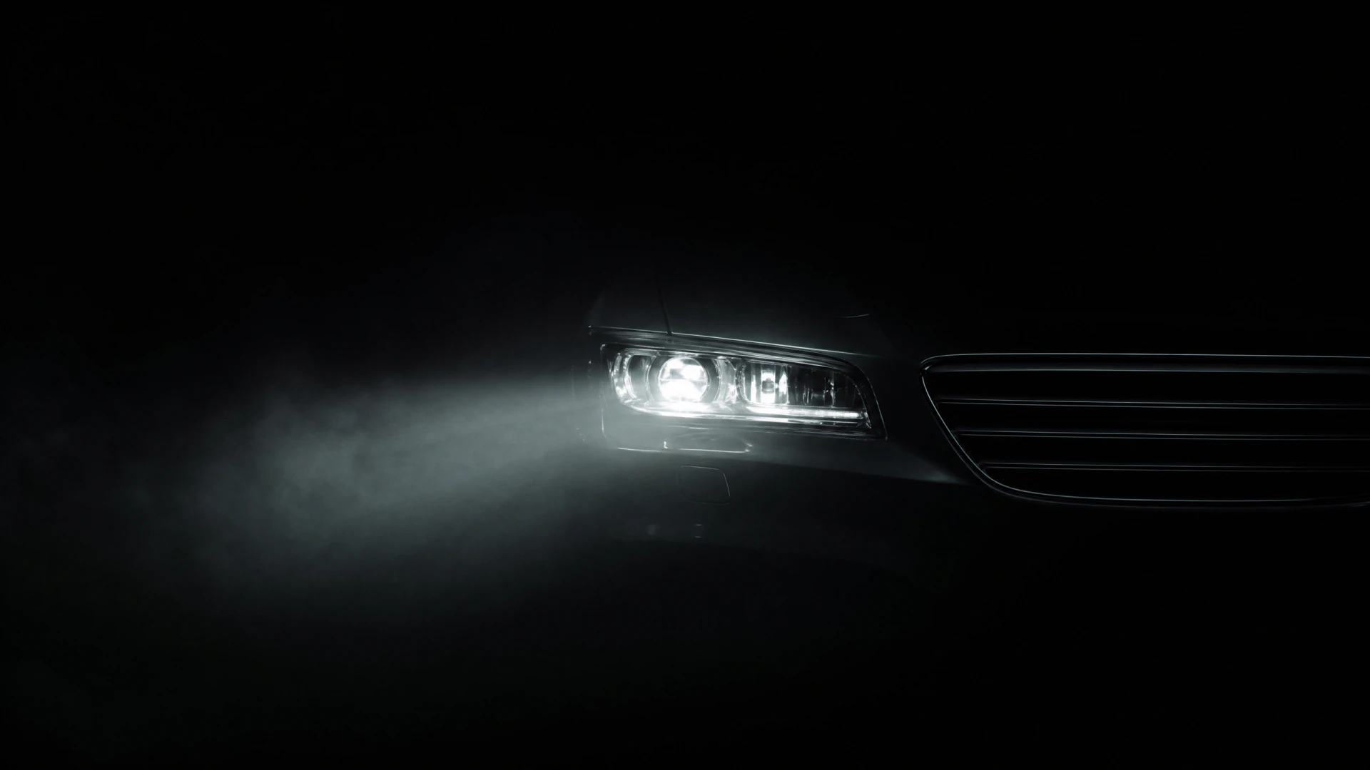 A car headlight glowing in darkness, with light scattering through low-lying mist in front of the vehicle.